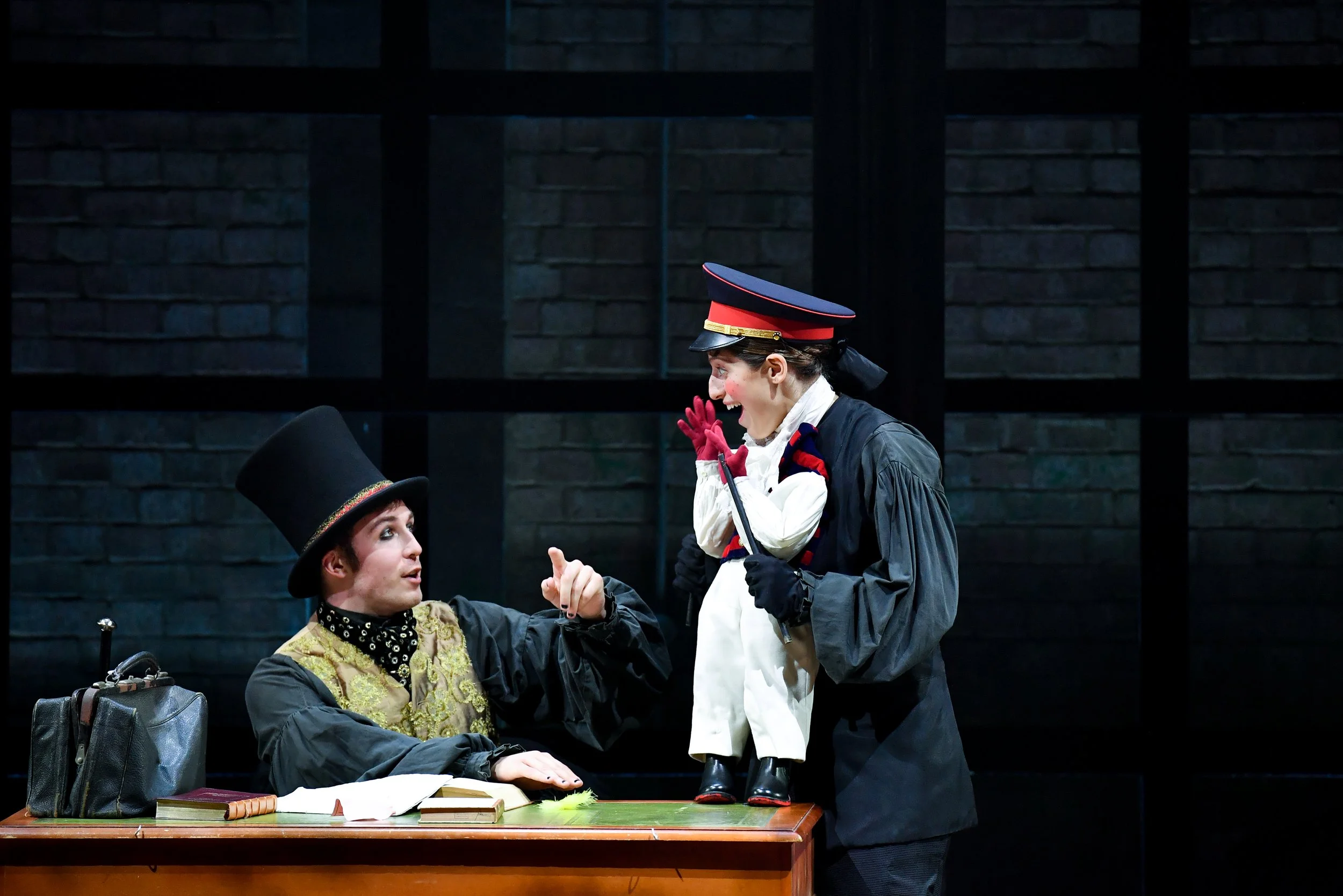 Two actors on stage, one sitting at a desk wearing a tall black hat, and the other standing with a military-style cap, holding a doll dressed in white. The person at the desk appears to be making a point, while the other is smiling and engaging with 
