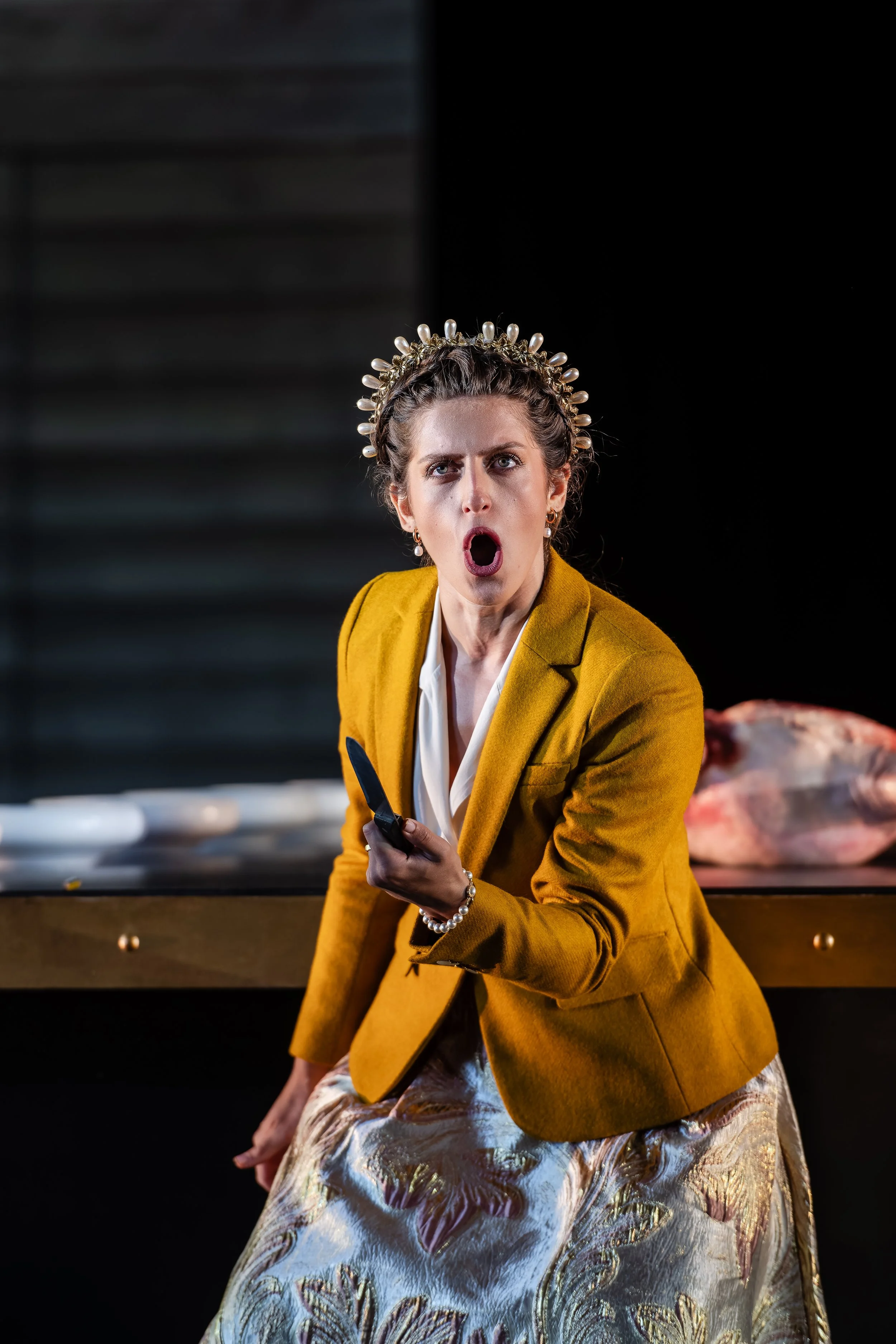 A woman with an angry expression, wearing a yellow blazer, a pearl necklace, and a decorative headpiece, holding a knife in a kitchen with a large fish on the counter.