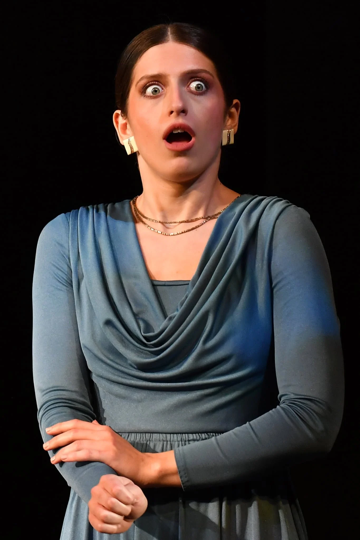 A woman with dark hair wearing a blue dress and gold jewelry, displaying a shocked or surprised expression.