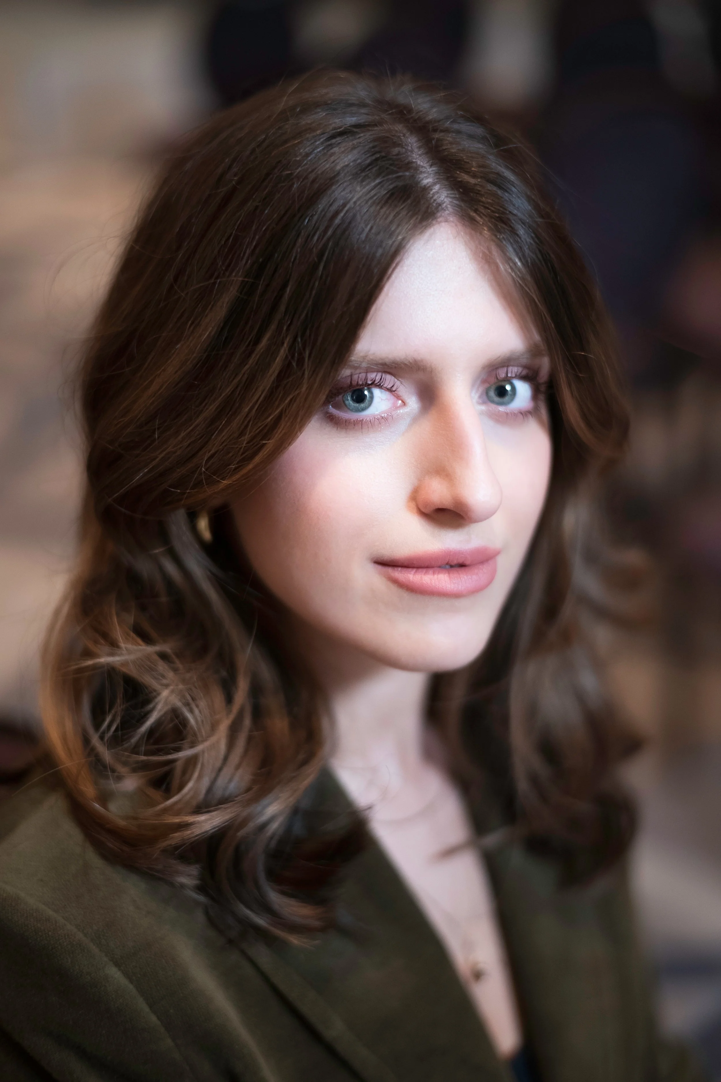 Close-up portrait of a woman with long brown hair and blue eyes, wearing a dark green blazer.