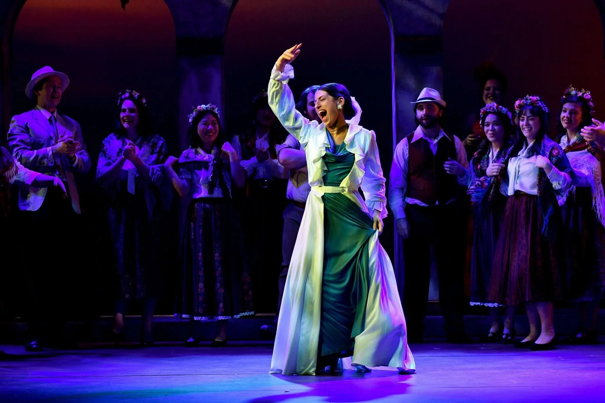 A musical theater performance with a cast on stage, featuring a woman in a green dress and white jacket dancing in the center, surrounded by other performers in period costumes.