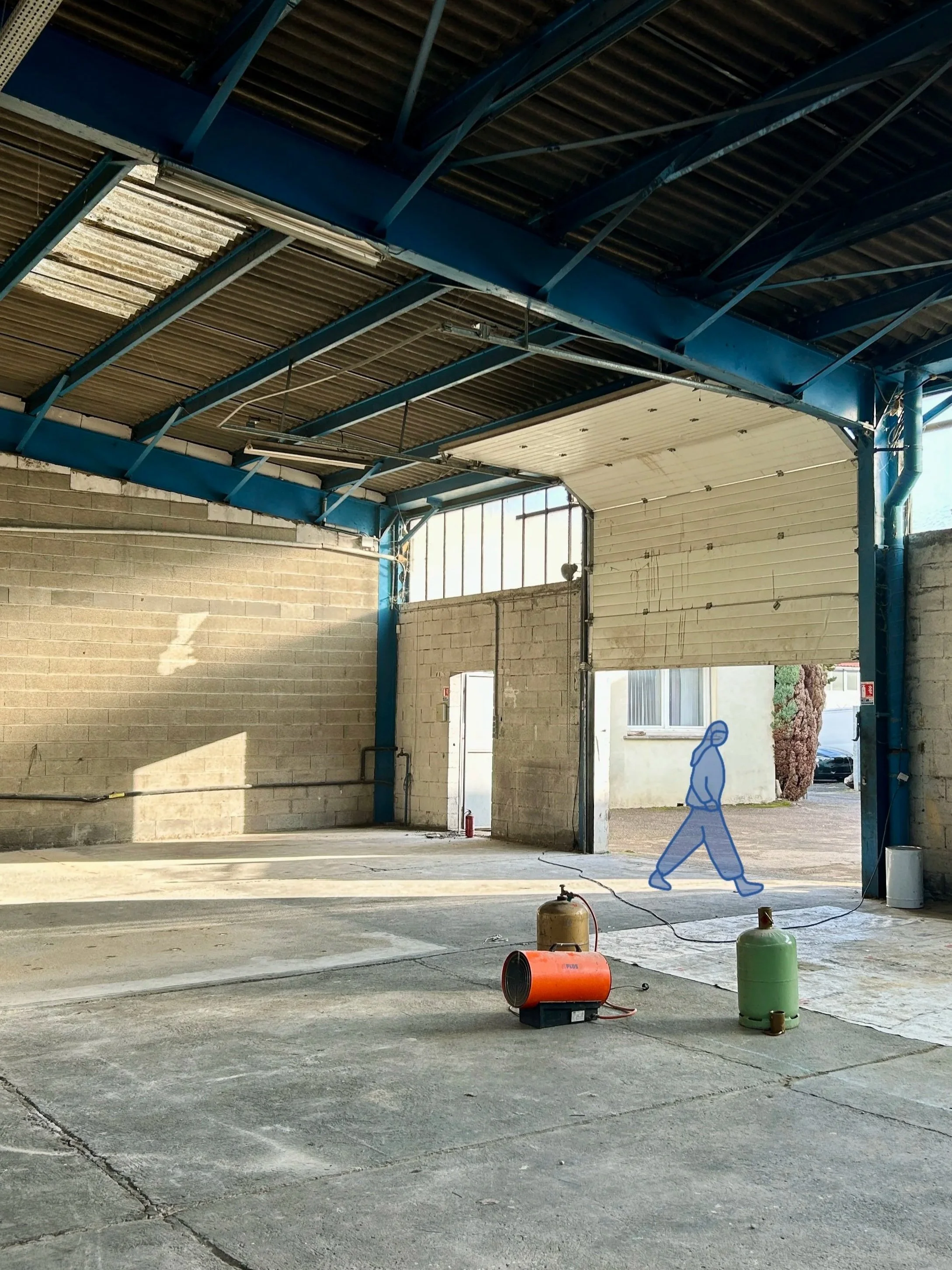 Intérieur d'un entrepôt ou d'un atelier en cours de construction ou de rénovation, avec un sol en béton et des murs en briques, un plafond en métal, et des équipements de chantier tels qu'un générateur électrique, des cylindres de gaz, et un câble électrique.