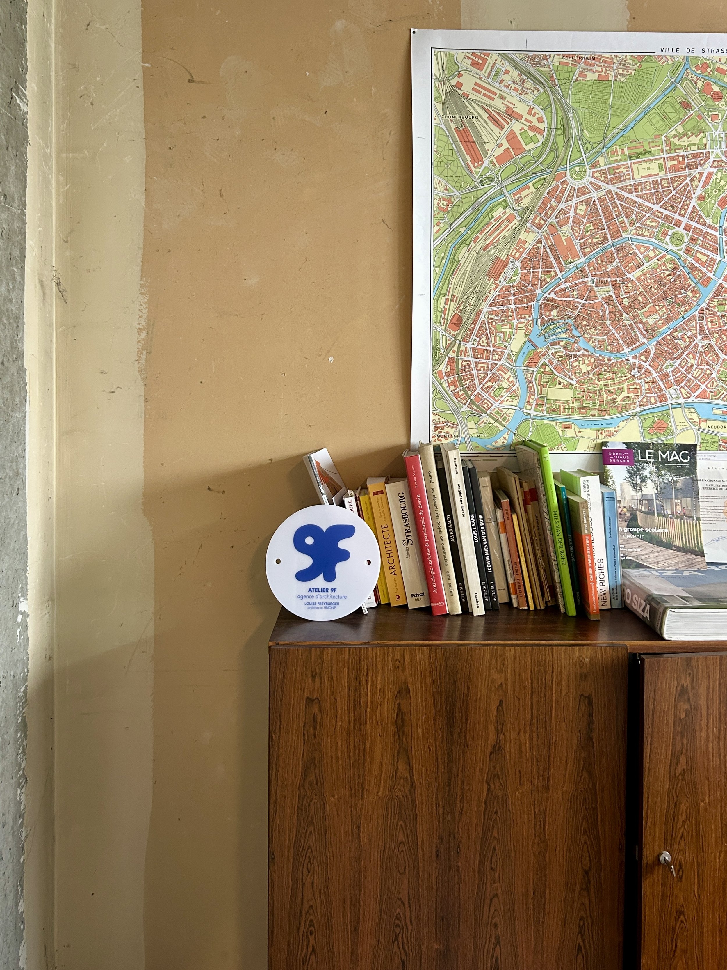 Un meuble en bois avec des livres empilés dessus, une carte de Strasbourg au mur, et un panneau indiquant Atelier 9F, dans une pièce avec un mur beige et un sol en béton, intérieur de l'agence d'architecture.