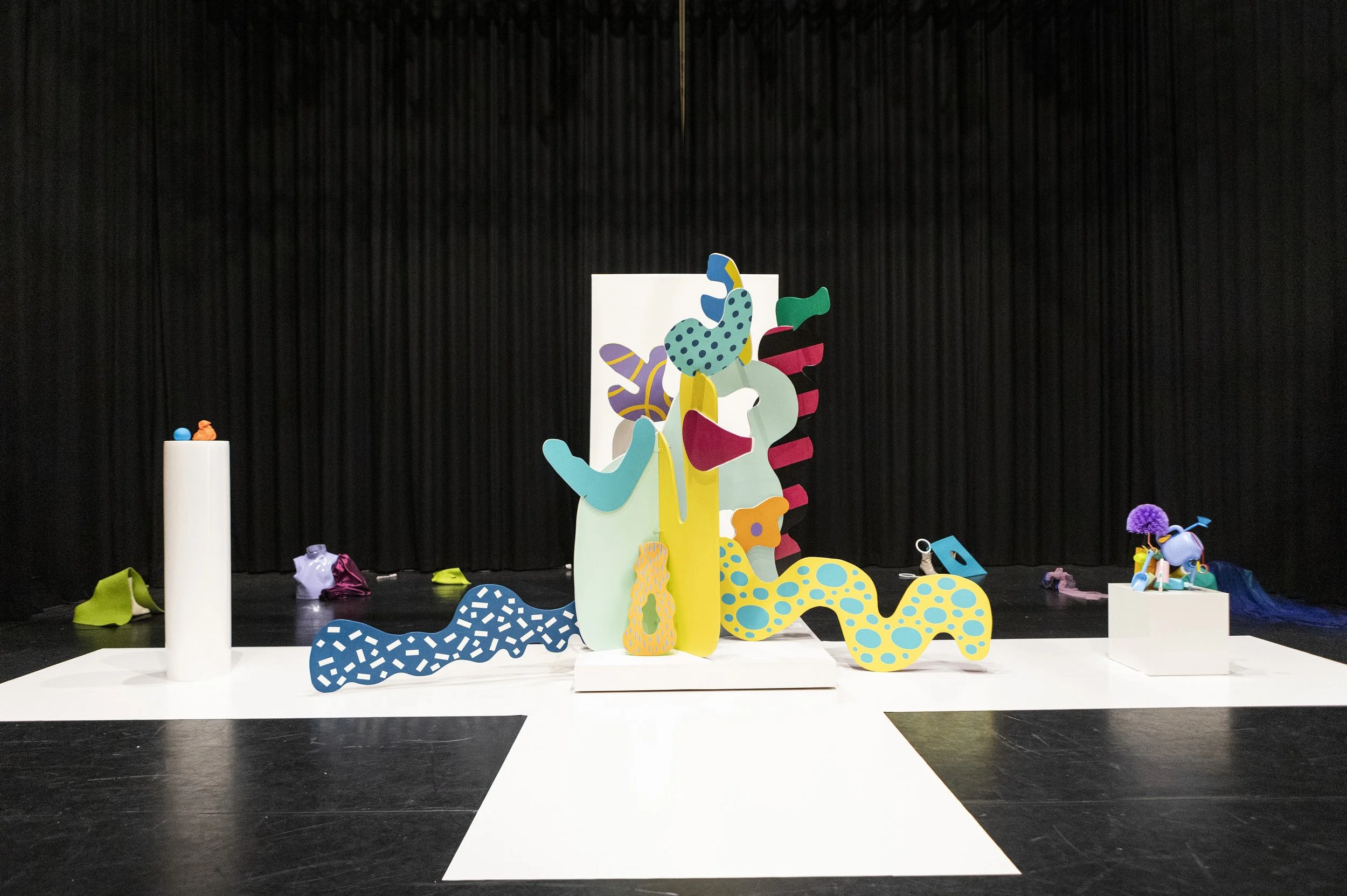 Kleurrijke, abstracte sculpturen en objecten op een podium met een zwarte achtergrond, mogelijk onderdeel van een tentoonstelling of kunstinstallatie.