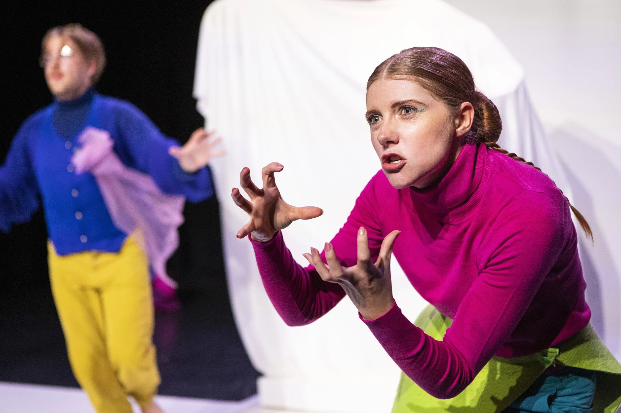 Vrouw in roze trui en groene broek in theaterstijl, die expressief met handen en gezicht communiceert. In de achtergrond zijn twee mensen zichtbaar, een in blauw jasje en gele broek, de ander onduidelijk uitvergroot.
