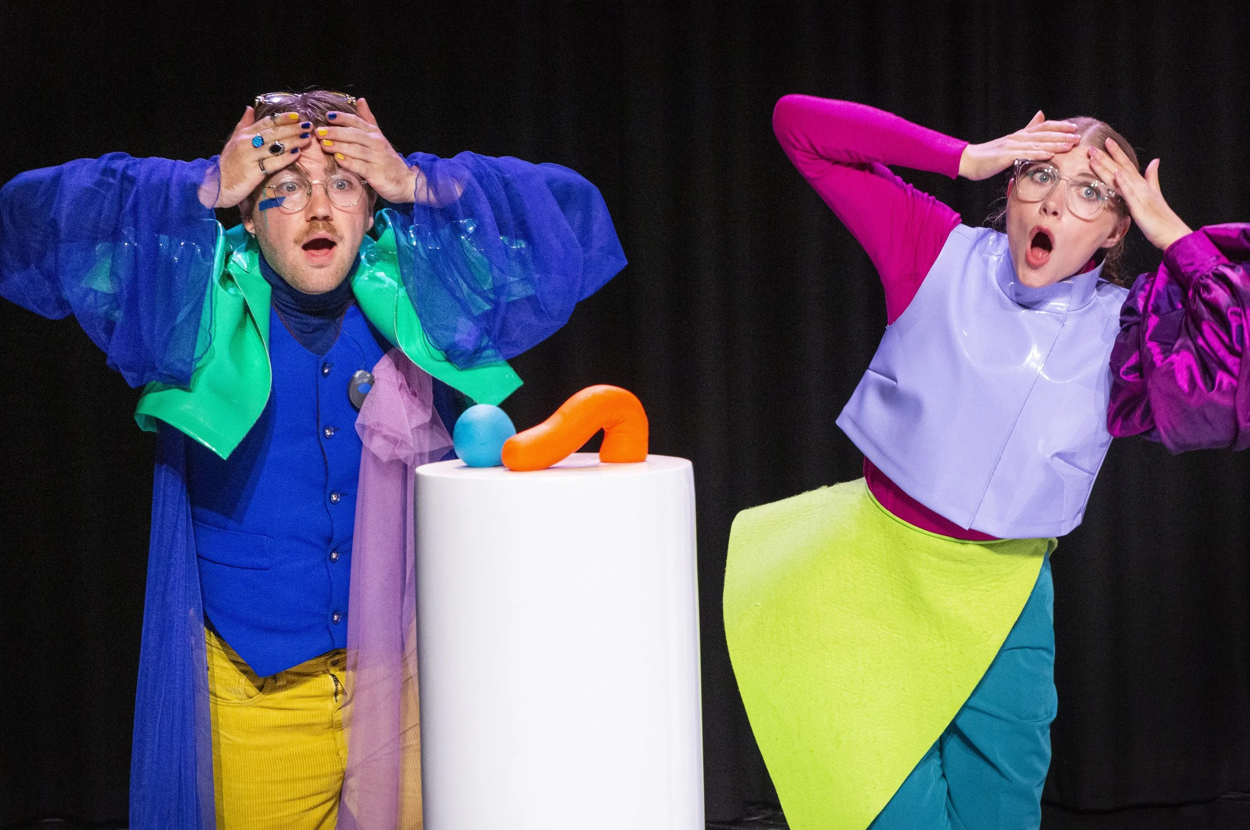 Twee mensen gekleed in felgekleurde outfits met handen op hun hoofd kijken verbaasd naar voorwerpen op een witte tafel.