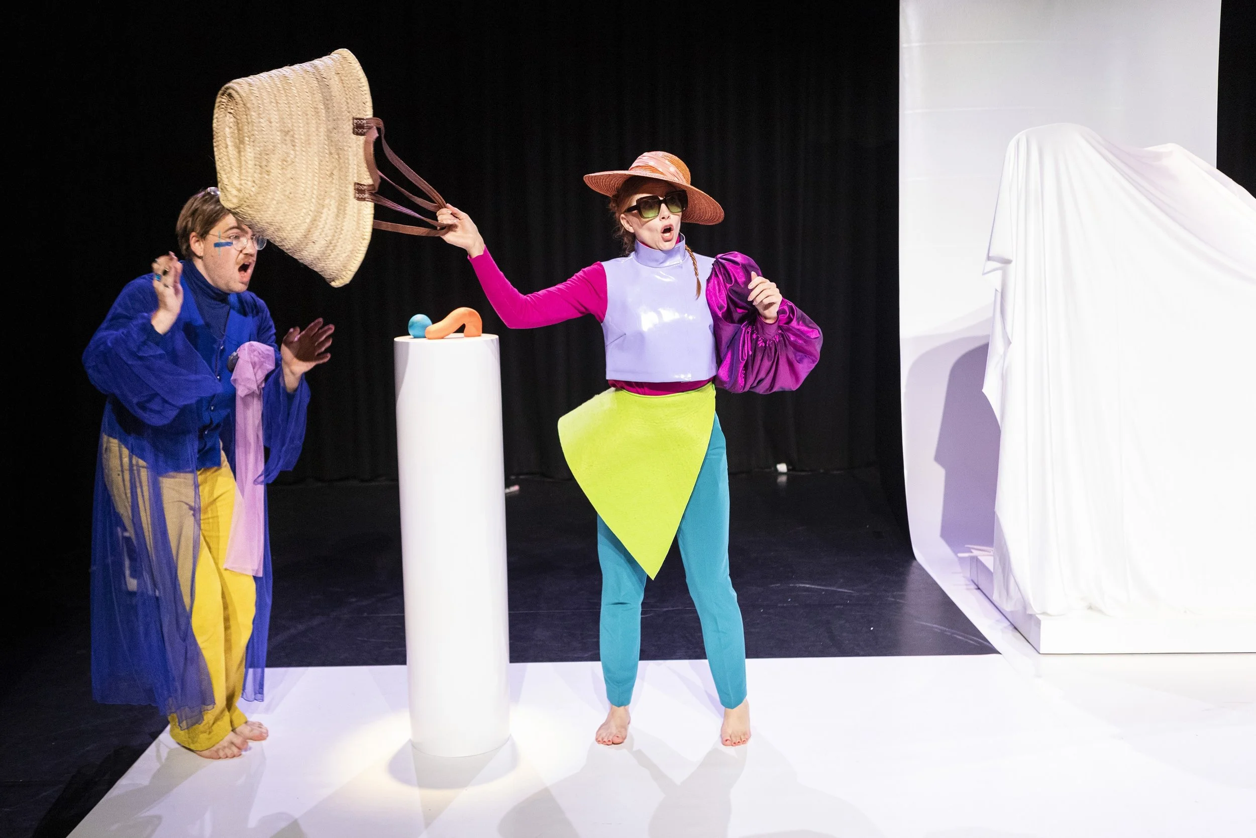 Twee acteurs op een podium doen een toneelstuk met kleurrijke kostuums. Een acteur houdt een groot rieten tas omhoog terwijl de andere met een grote hoed en zonnebril terugschreeuwt.