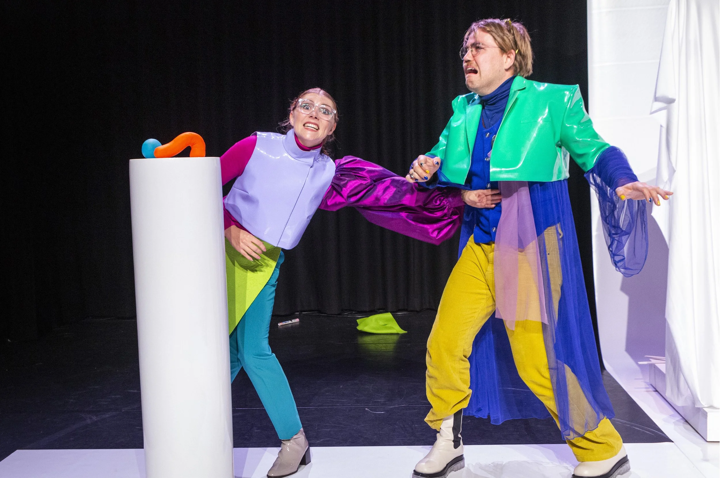 Twee mensen in kleurrijke outfits op een podium, een vrouw met een glazen muts en een man met een groene en paarse jas, betrokken in een dramatische scène.