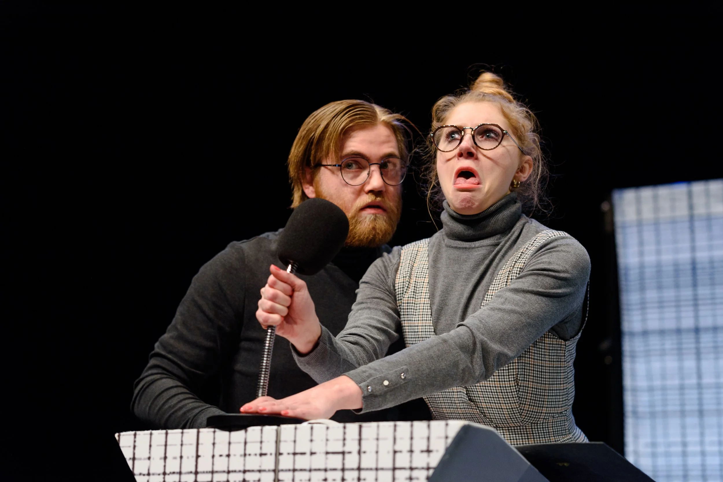 Twee mensen, een man met een baard en een vrouw met een knot in haar haar, dragen bril en maken een handdruk, terwijl ze naar links kijken. Ze staan op een podium met een zwarte achtergrond.