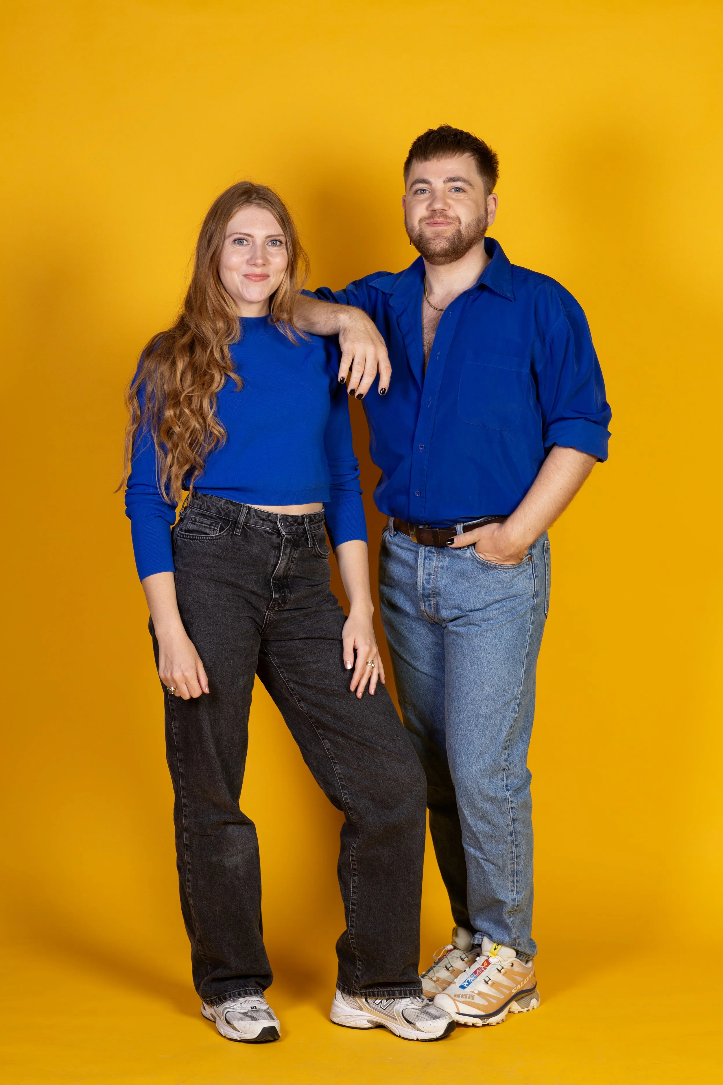 Twee jonge mensen staan samen tegen een geel achtergrond, de vrouw heeft lang rood haar en draagt een blauw shirt en zwarte broek, de man heeft kort bruin haar en draagt een blauw overhemd en blauwe jeans.