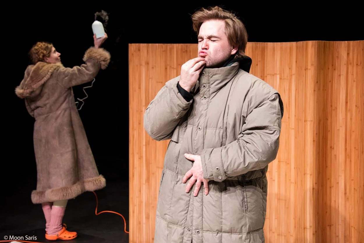 Een man in een beige winterjas met gesloten ogen en een peinzende uitdrukking, met een houten wand op de achtergrond. Op de achtergrond een vrouw in een lange bontkleurige jas die een apparaat vasthoudt, mogelijk een luchtbevochtiger of geurverspreider, met draden ernaast.