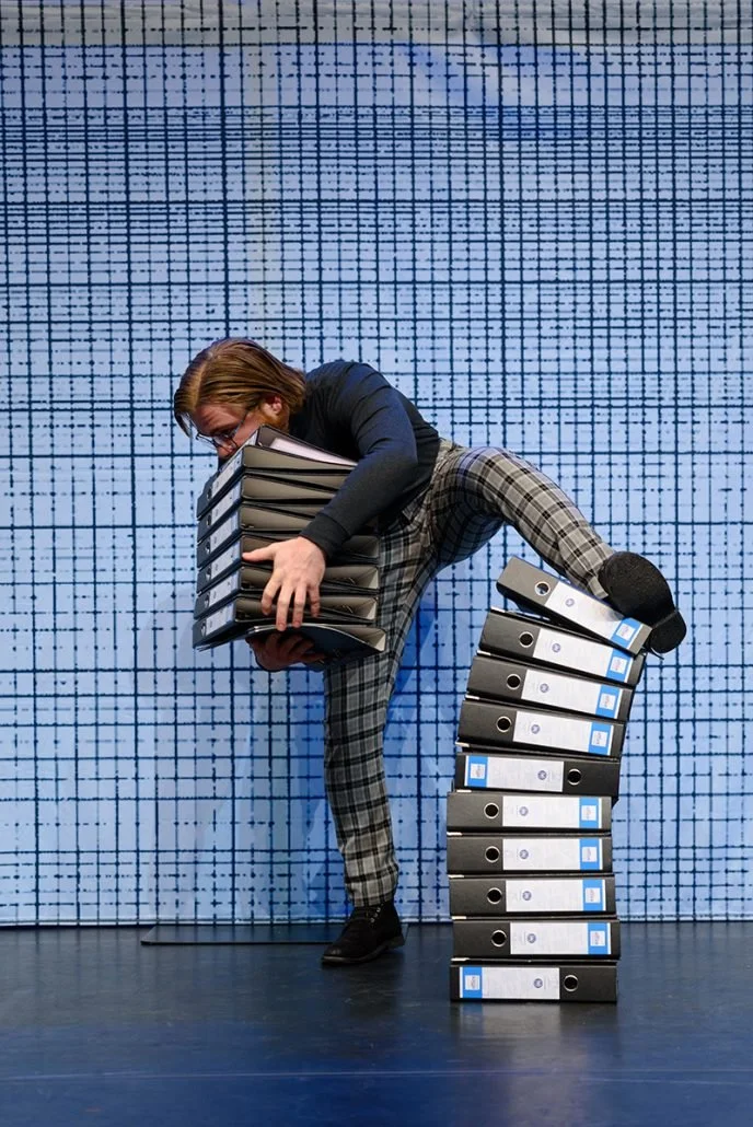 Man met lang haar, bril, donker jack en geblokte broek die probeert een stapel bestanden te dragen, terwijl hij balanceert op een rij bestanddozen tegen een blauwe, gemaaide achtergrond.