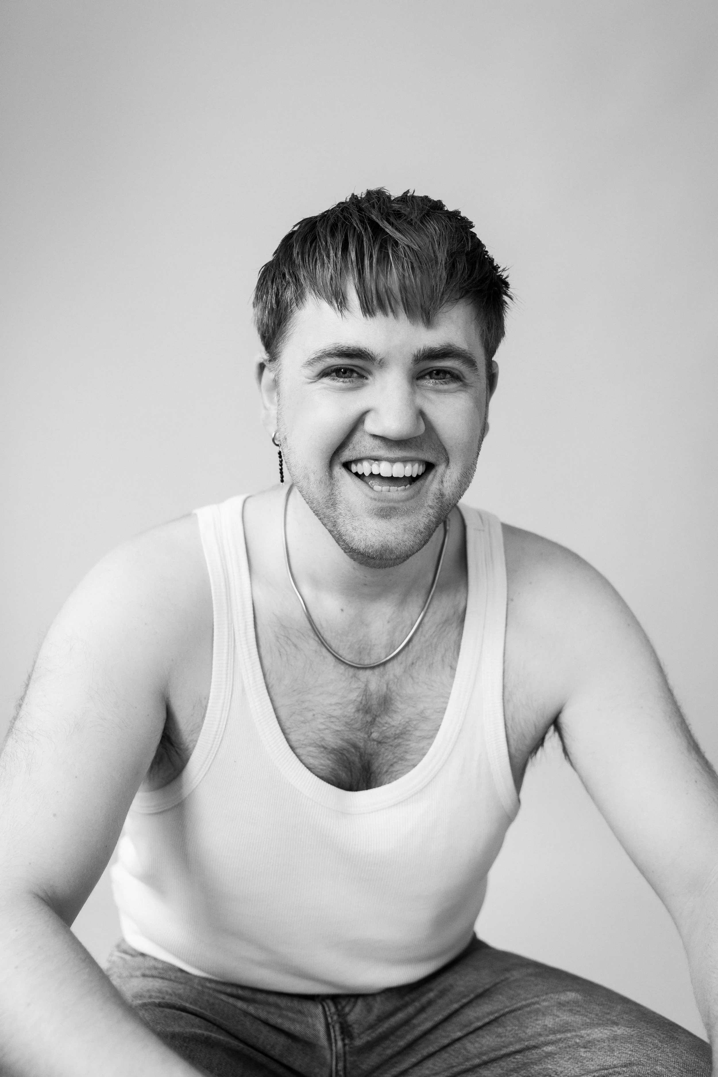 Lachende jonge man met kort haar, een oorring, in een witte tanktop en grijze broek, zittend tegen een effen muur.