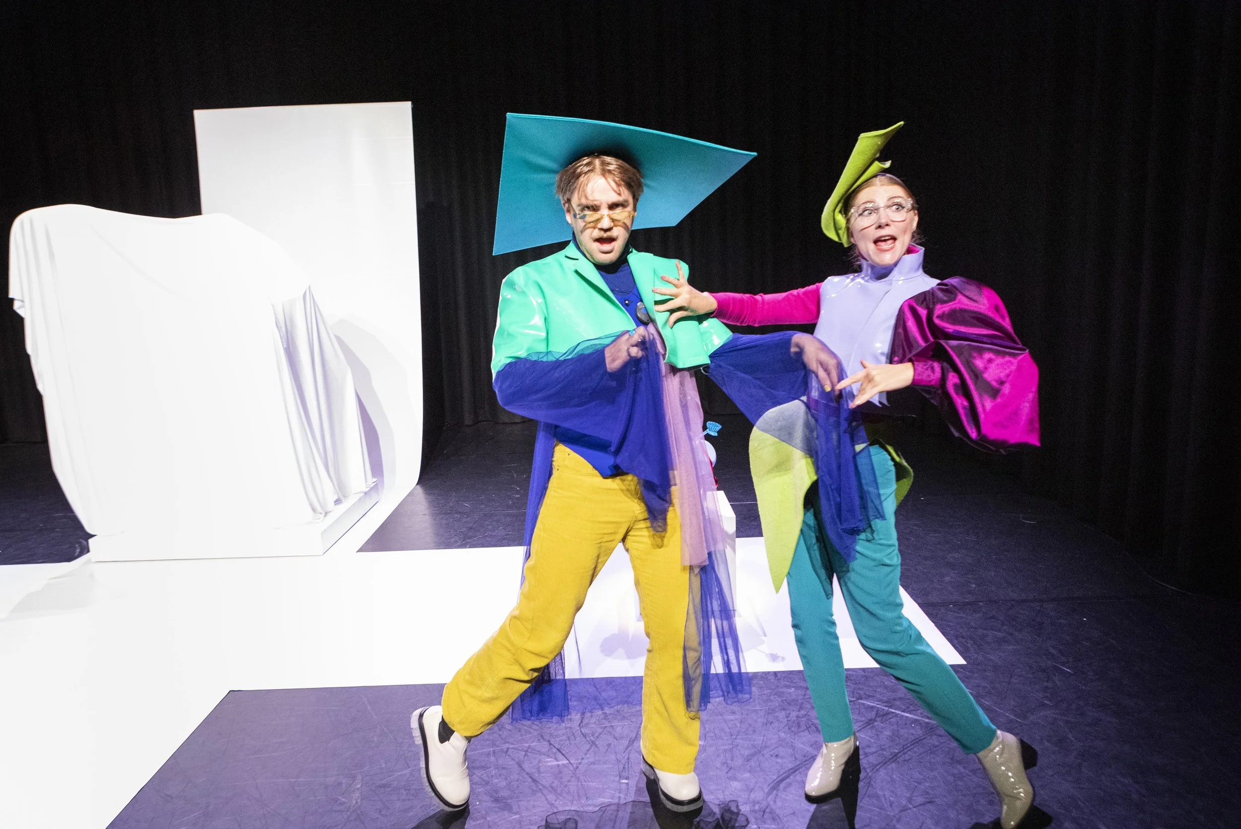 Twee acteurs in kleurrijke kostuums op een podium, onder een zwart gordijn, met een witte sculptuur links op de achtergrond.