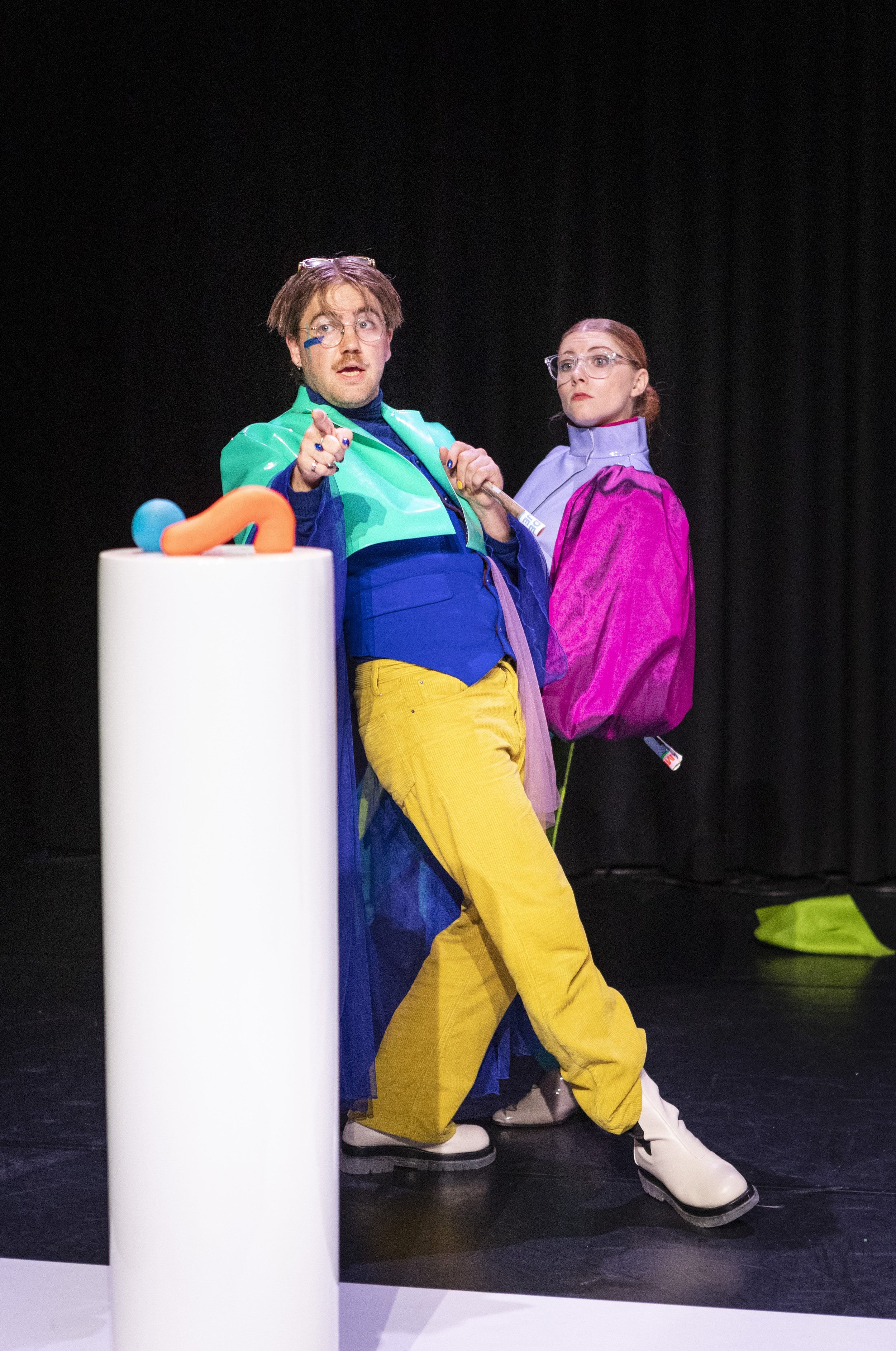 Twee mensen in kleurrijke outfits op een podium, met een zwarte achtergrond, terwijl ze in beweging lijken te zijn.