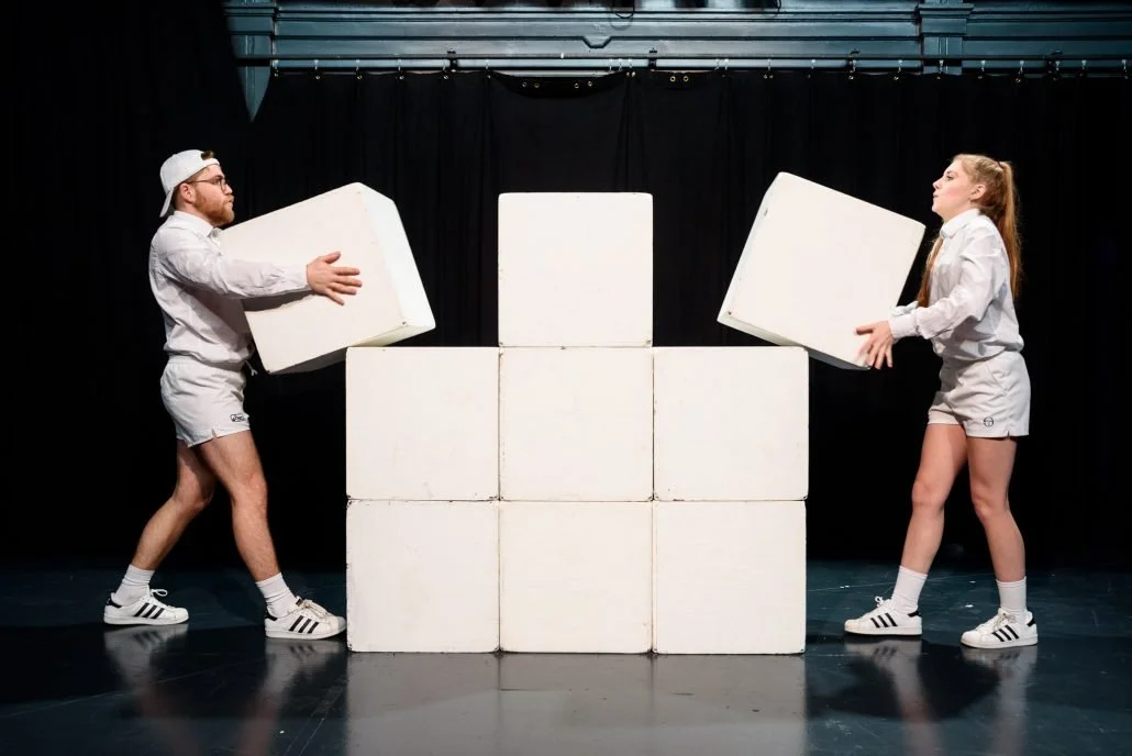 Twee mensen, een man en een vrouw, bouwen met grote witte blokken op een podium met zwarte achtergrond.