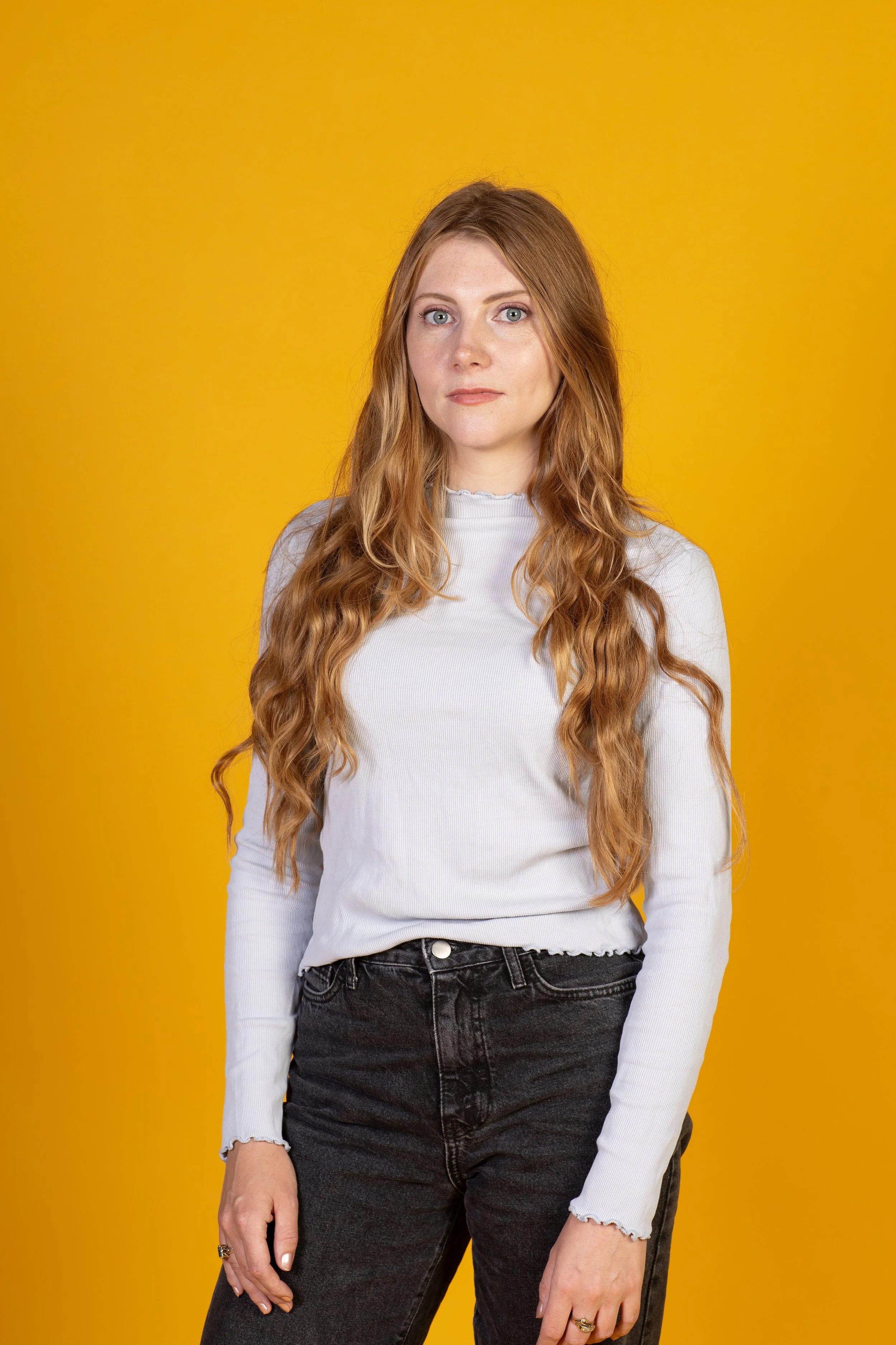 Een portret van een vrouw met lang, golvend rood haar, blauwe ogen, een lichte huid en een neutrale uitdrukking, gedragen in een witte langarm blouse en zwart jeans, staande voor een gele achtergrond.