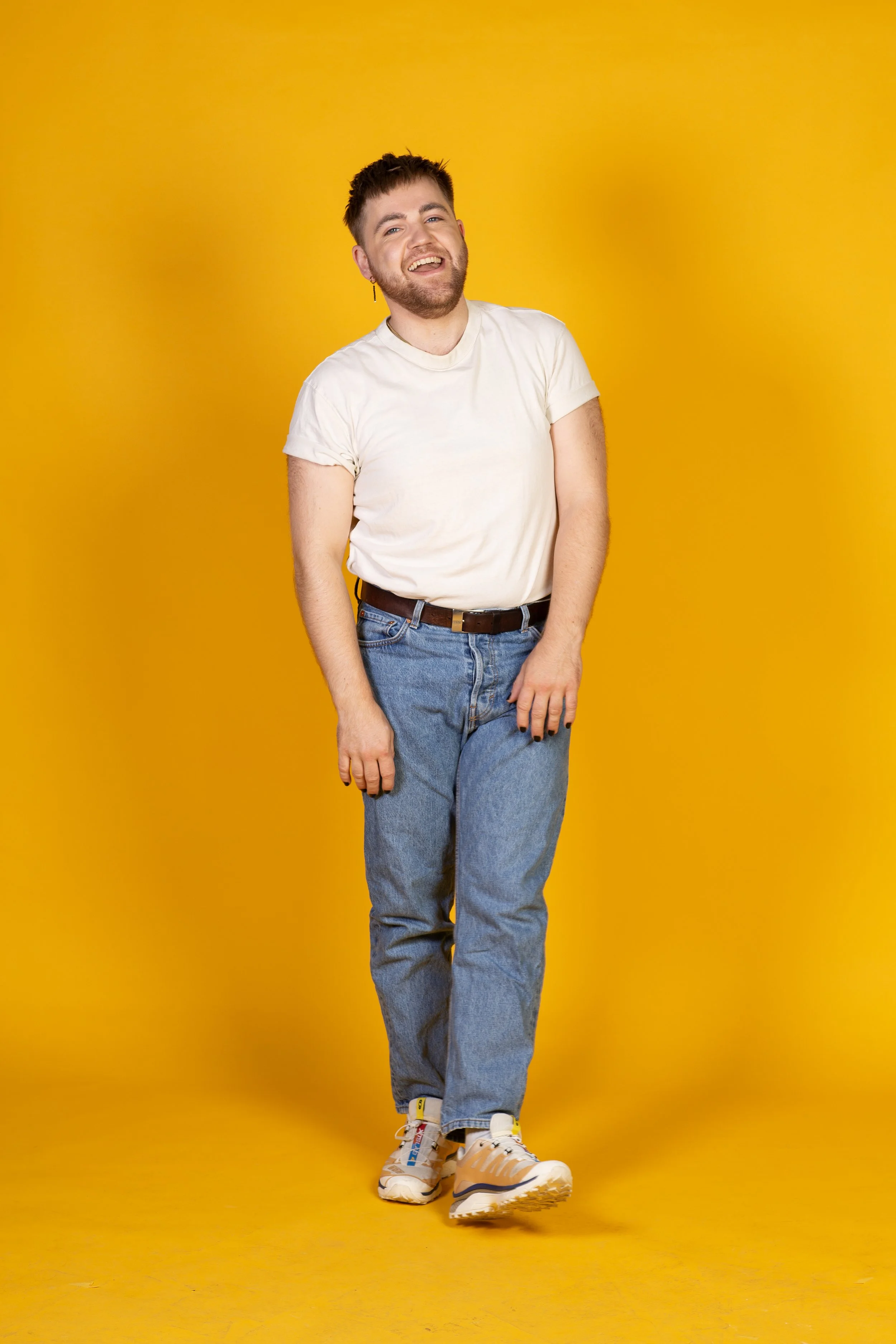 Man draagt een witte T-shirt, blauwe jeans en sneakers, staat lachend voor een gele achtergrond.
