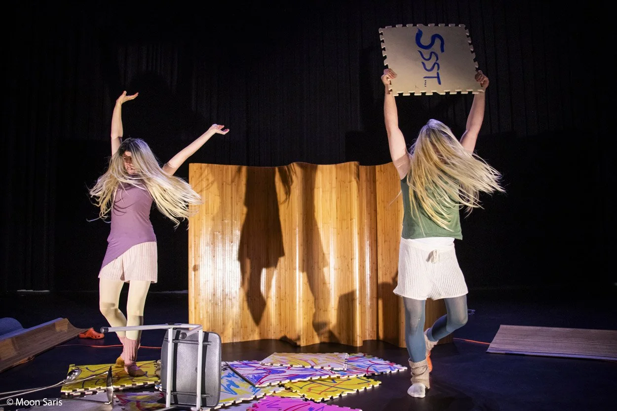 Twee jonge meisjes dansen op een podium, een van hen houdt een bord met de tekst 'SISS' omhoog. Ze dragen kleurrijke kleding en lijken plezier te hebben. De achtergrond is zwart en er liggen puzzelstukken op de vloer.
