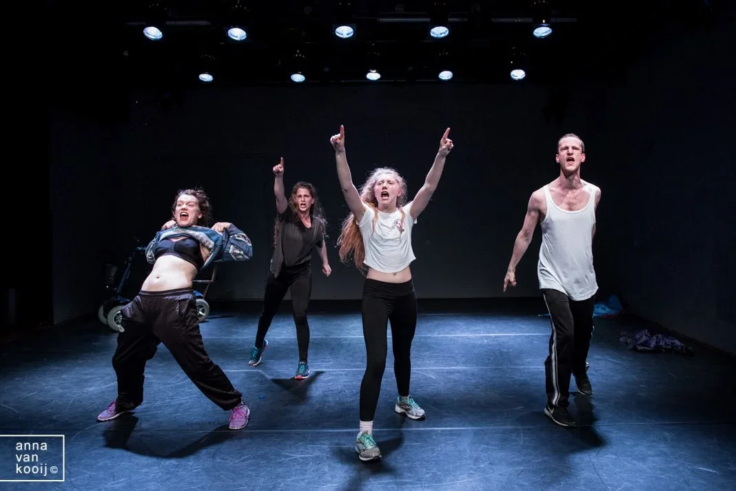 Vier mensen oefenen een theatervoorstelling op een donkere podium, met enkele lichte spots boven hen.