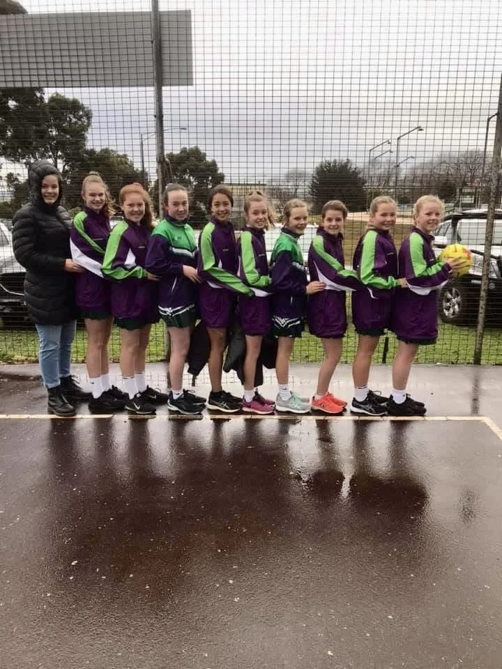 A girls' sports team standing in a line on a wet outdoor court, some holding a yellow netball, with a chain-link fence and trees in the background.