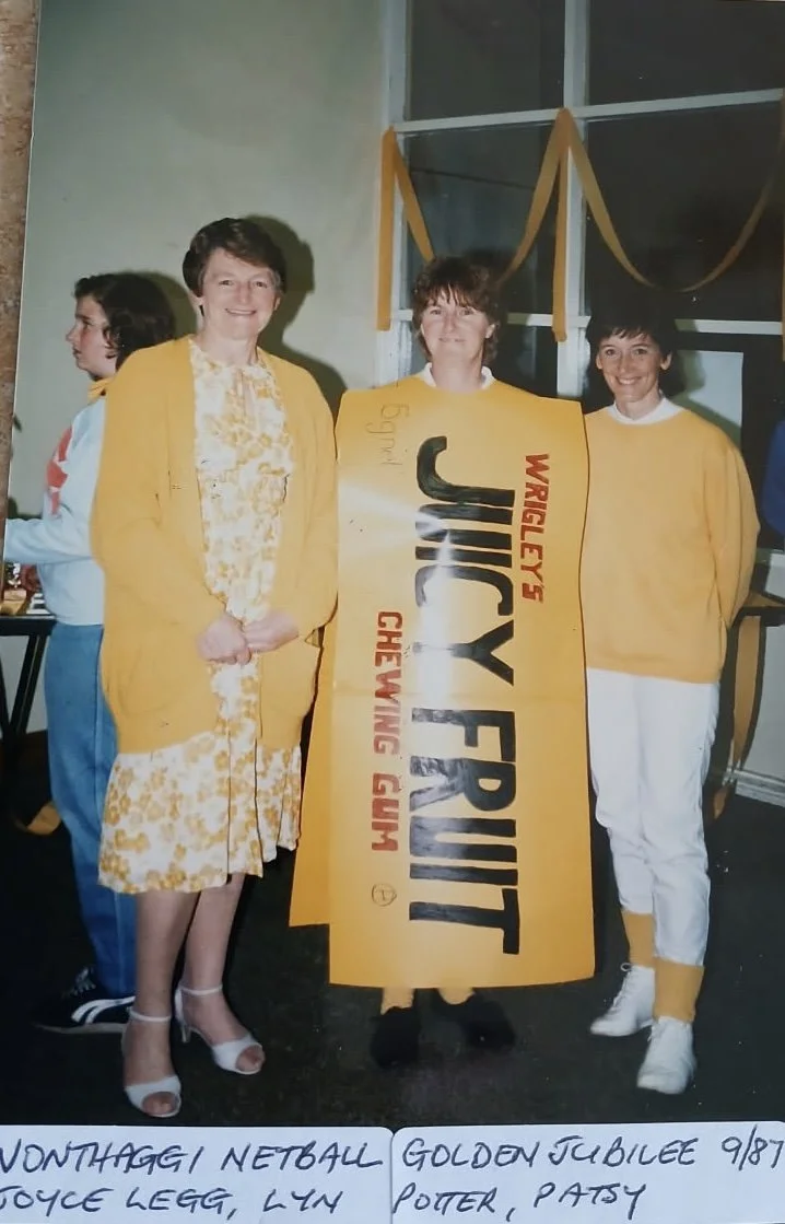 Group of three women standing indoors, with two of them wearing matching yellow sweaters and a yellow dress , and the third holding a large sign for Jucy Fruit bubble gum. Commemorating the grand jubillee. Decorations are visible in the background.