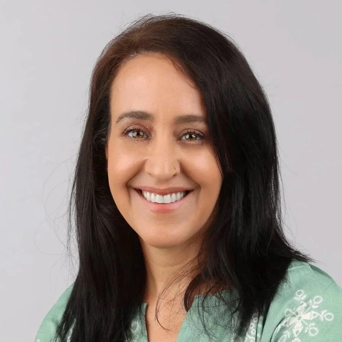Smiling woman with shoulder-length black hair, wearing a light green top with white embroidery, against a plain light gray background.