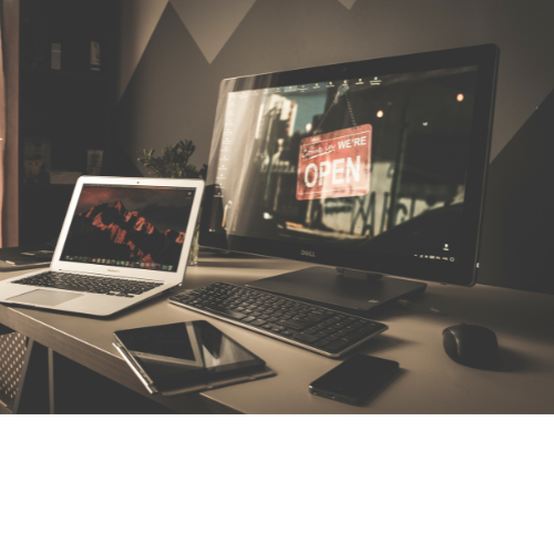 Desk setup with a large monitor displaying a neon 'We Are Open' sign, a laptop showing a red-lit cityscape, a tablet, a smartphone, a keyboard, and a mouse on a white desk in a modern office space.
