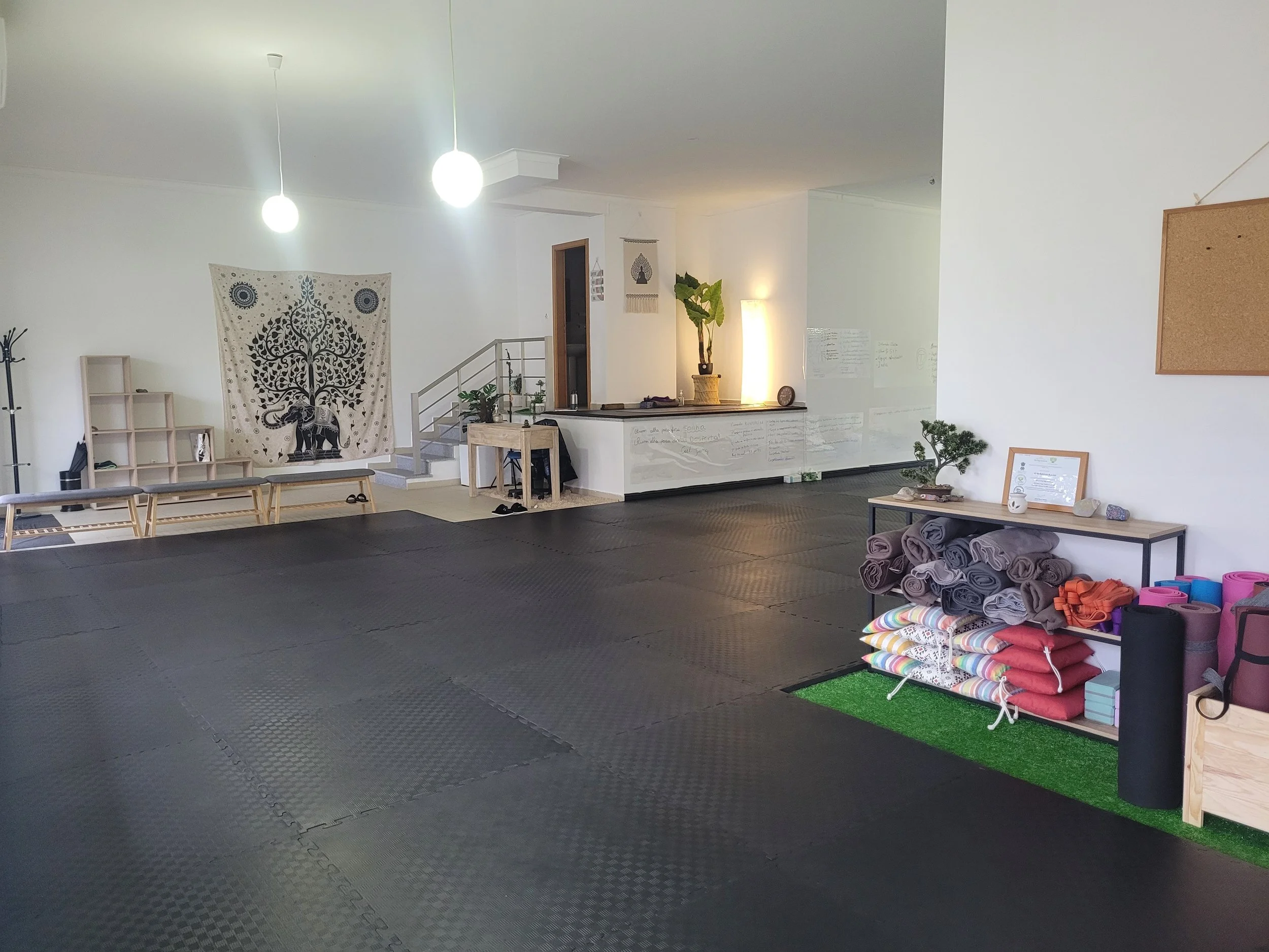 Estúdio de yoga com tapetes pretos no chão, uma prateleira com toalhas enroladas, uma pequena mesa com plantas e uma grande tapeçaria decorativa com um elefante e uma árvore na parede. Iluminação suave com duas luminárias pendentes no teto.