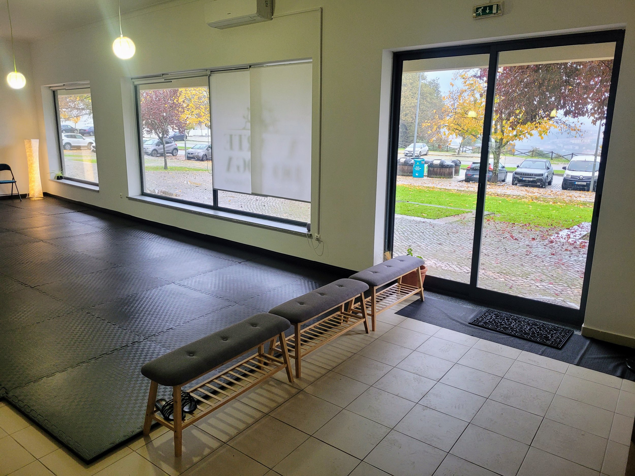 Interior de um estúdio de yoga com grandes janelas que mostram um parque de estacionamento exterior e árvores com folhagem de outono. A sala tem um piso acolchoado preto de um lado, um piso de azulejo do outro e bancos perto da porta.