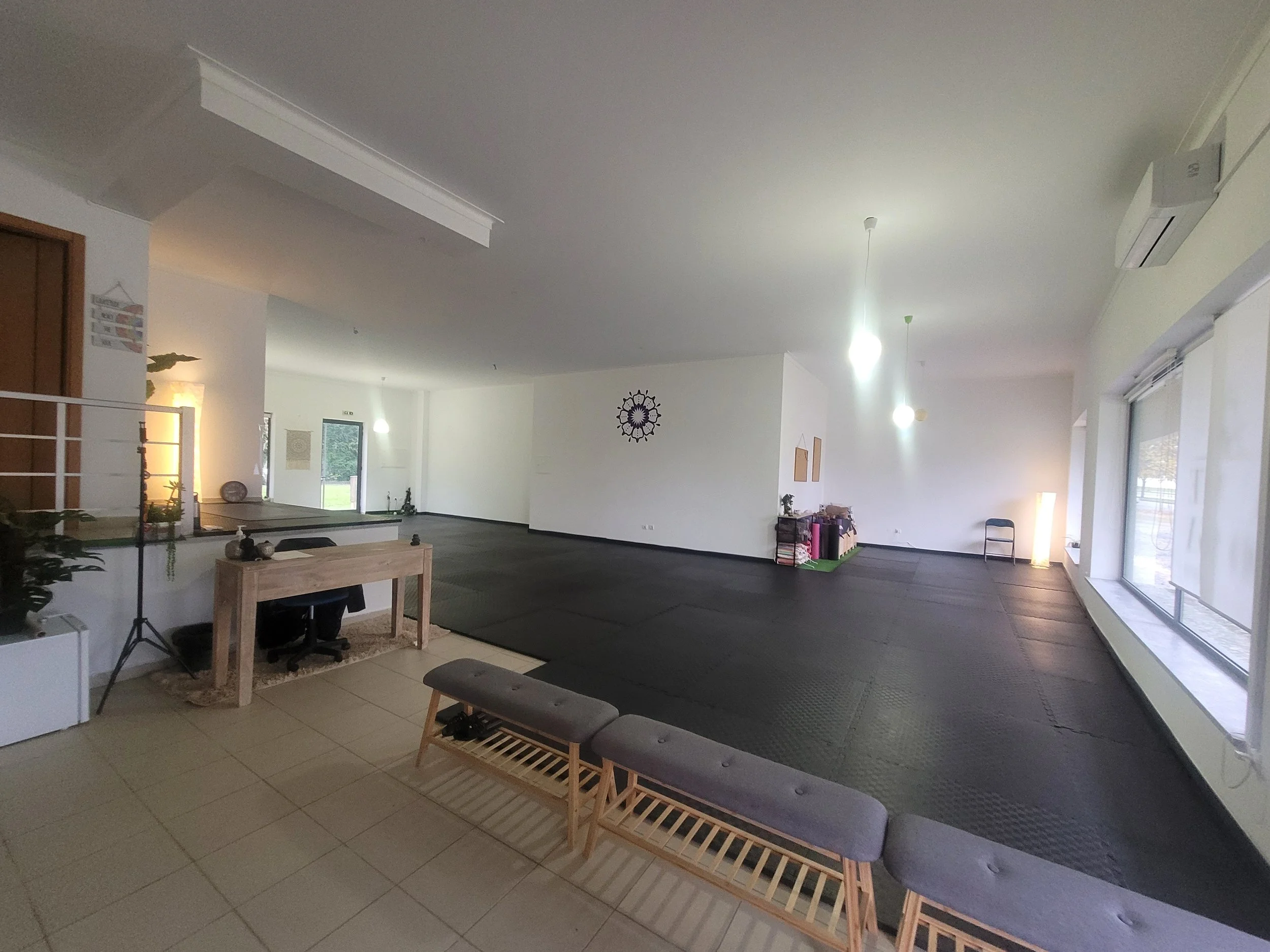 Um estúdio de yoga espaçoso e bem iluminado, com tapetes pretos no chão, uma pequena mesa com cadeira e decoração minimalista. Grandes janelas à direita permitem a entrada de luz natural.