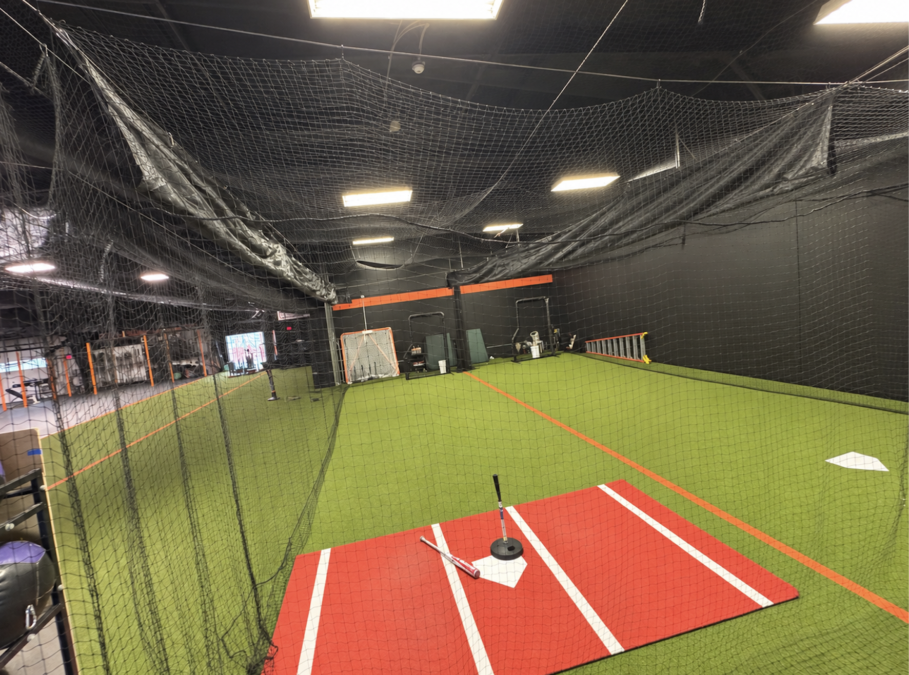 An indoor baseball or softball training facility with a green turf field, a red batting mat, and baseball equipment. The area is enclosed with black netting for batting practice.