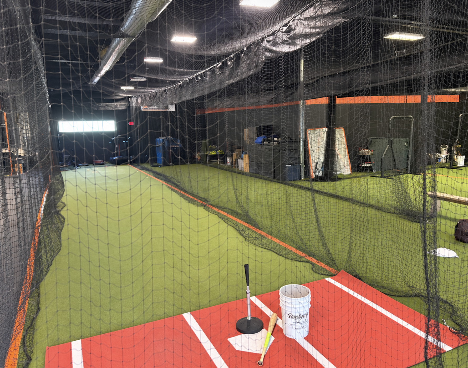 Indoor batting cage with artificial green turf and red and white batter's box markings, enclosed by black netting, with sports equipment and storage area in the background.