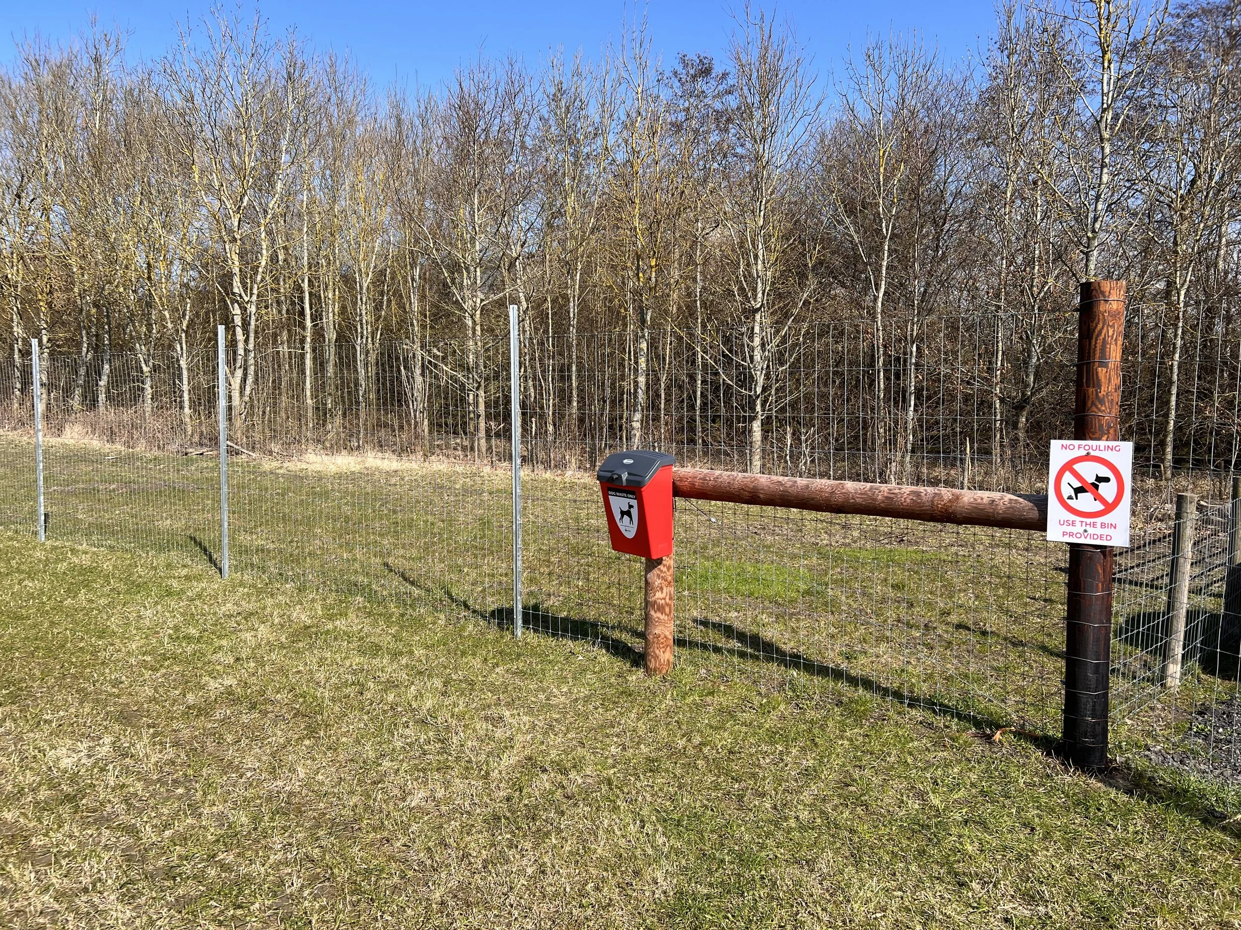 Fence surrounding a grassy area with a sign and a bin. The sign indicates no fouling and instructs to use the provided bin. The background features leafless trees and a clear blue sky.