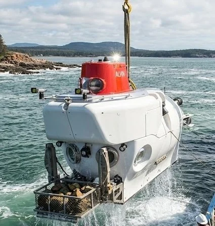 Autonomous underwater vehicle with a red top and white body, floating on water near a coastline with rocky shores and distant hills.