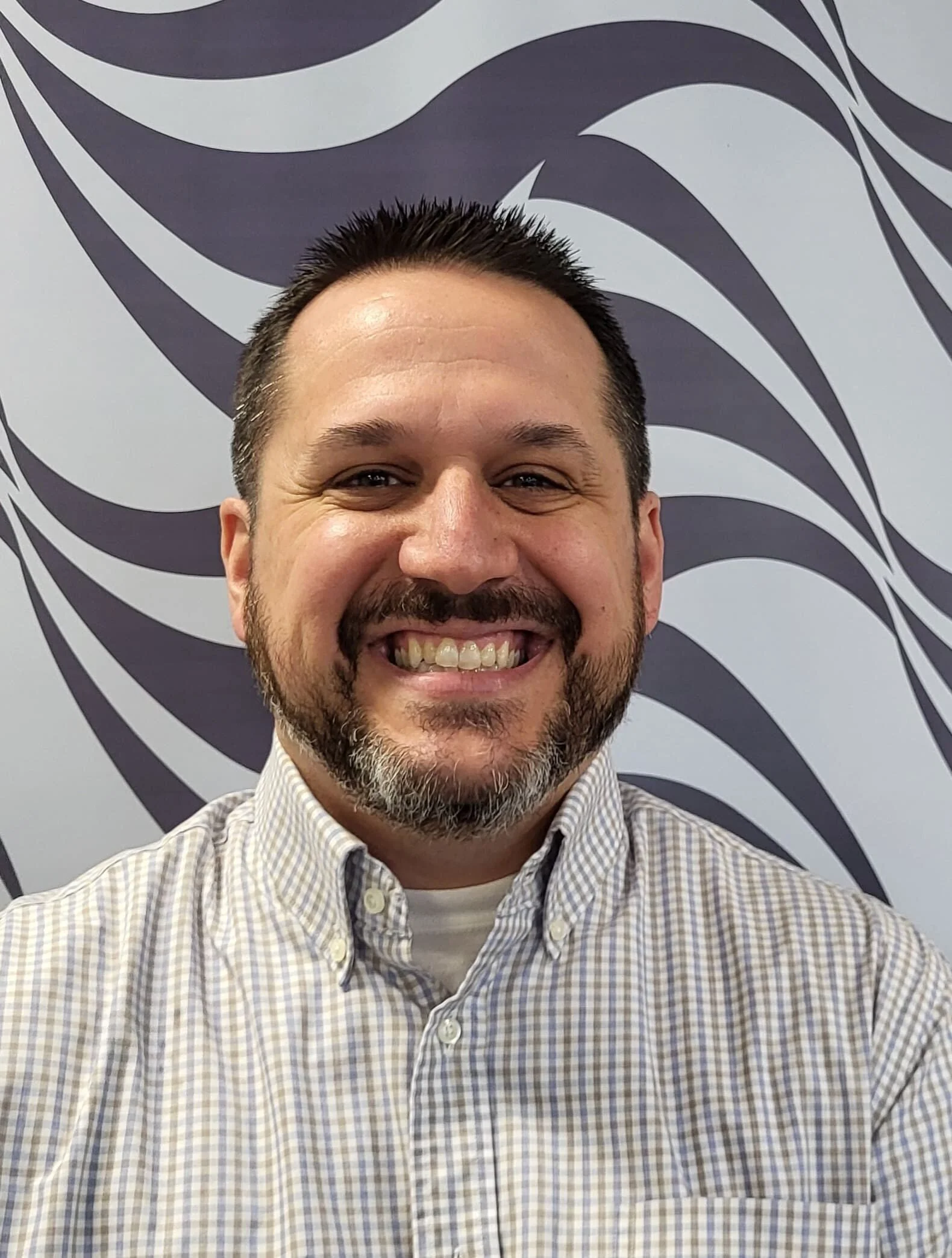 A smiling man with short dark hair and a beard, wearing a checkered button-up shirt, standing in front of a decorative, abstract black and white background.