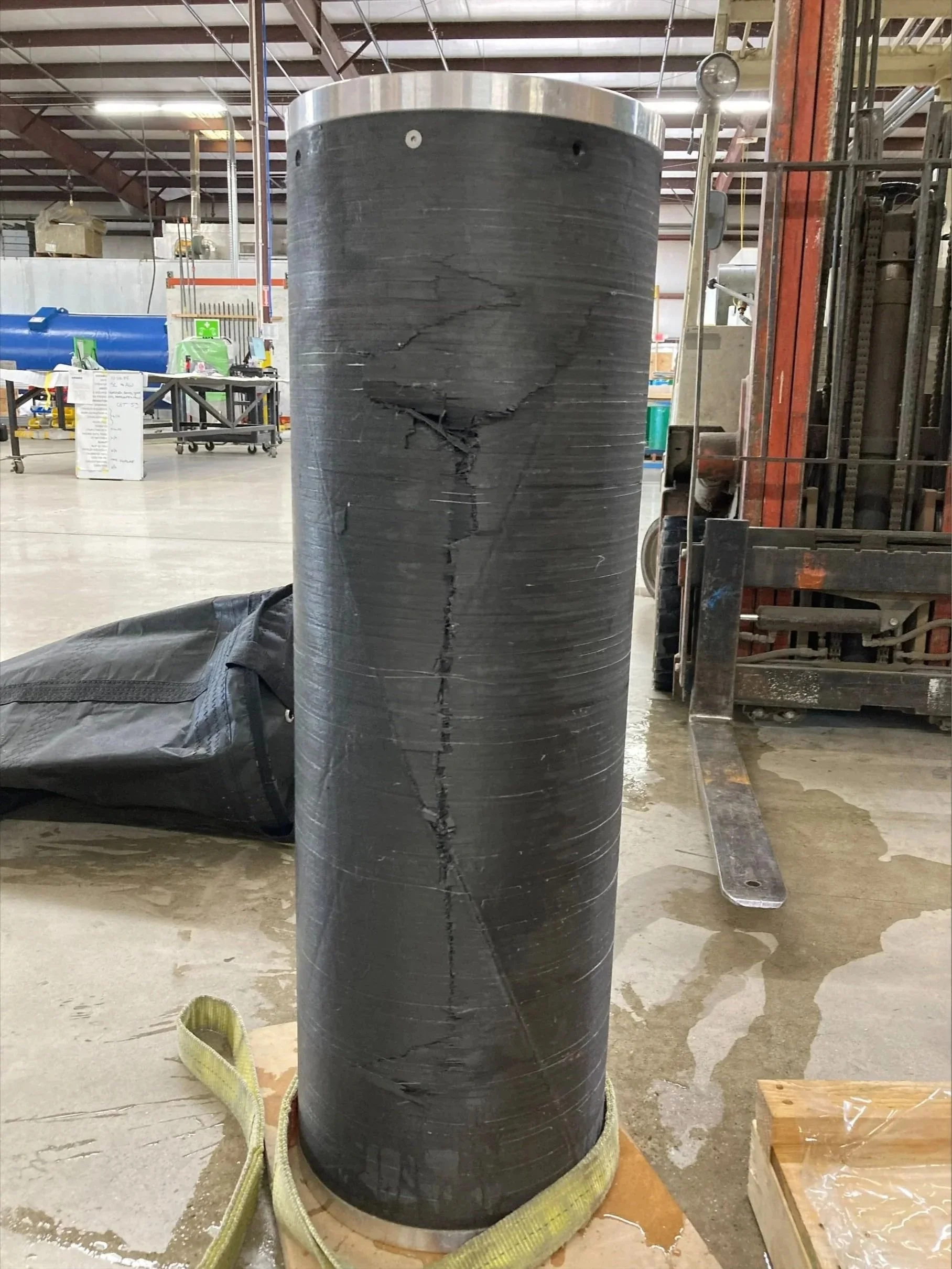 A large, black cylindrical object with visible cracks, standing upright on a wooden base in an industrial warehouse. The object appears damaged.