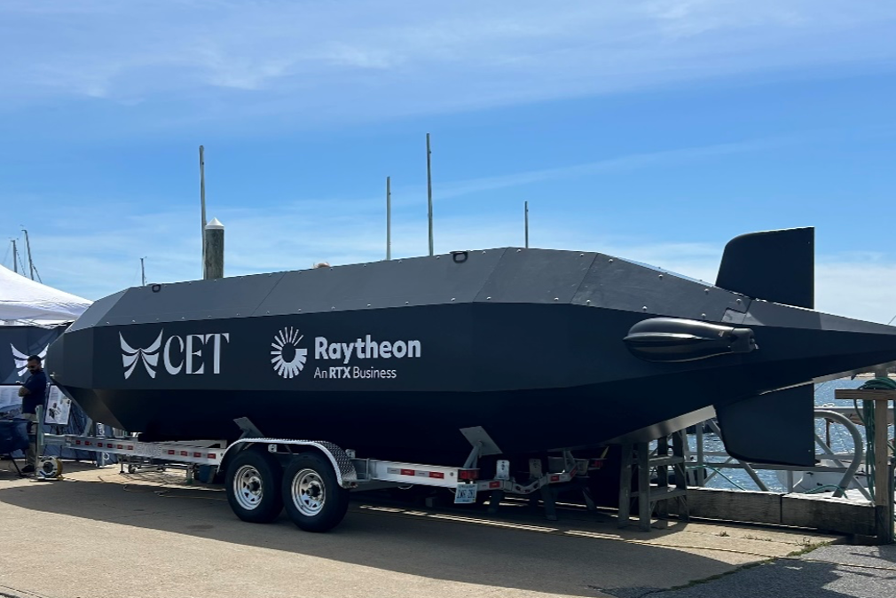 A large black underwater drone resembling a torpedo on a trailer, displaying logos for CET, Raytheon, and RTX Business, parked near a dock with water and boats in the background.