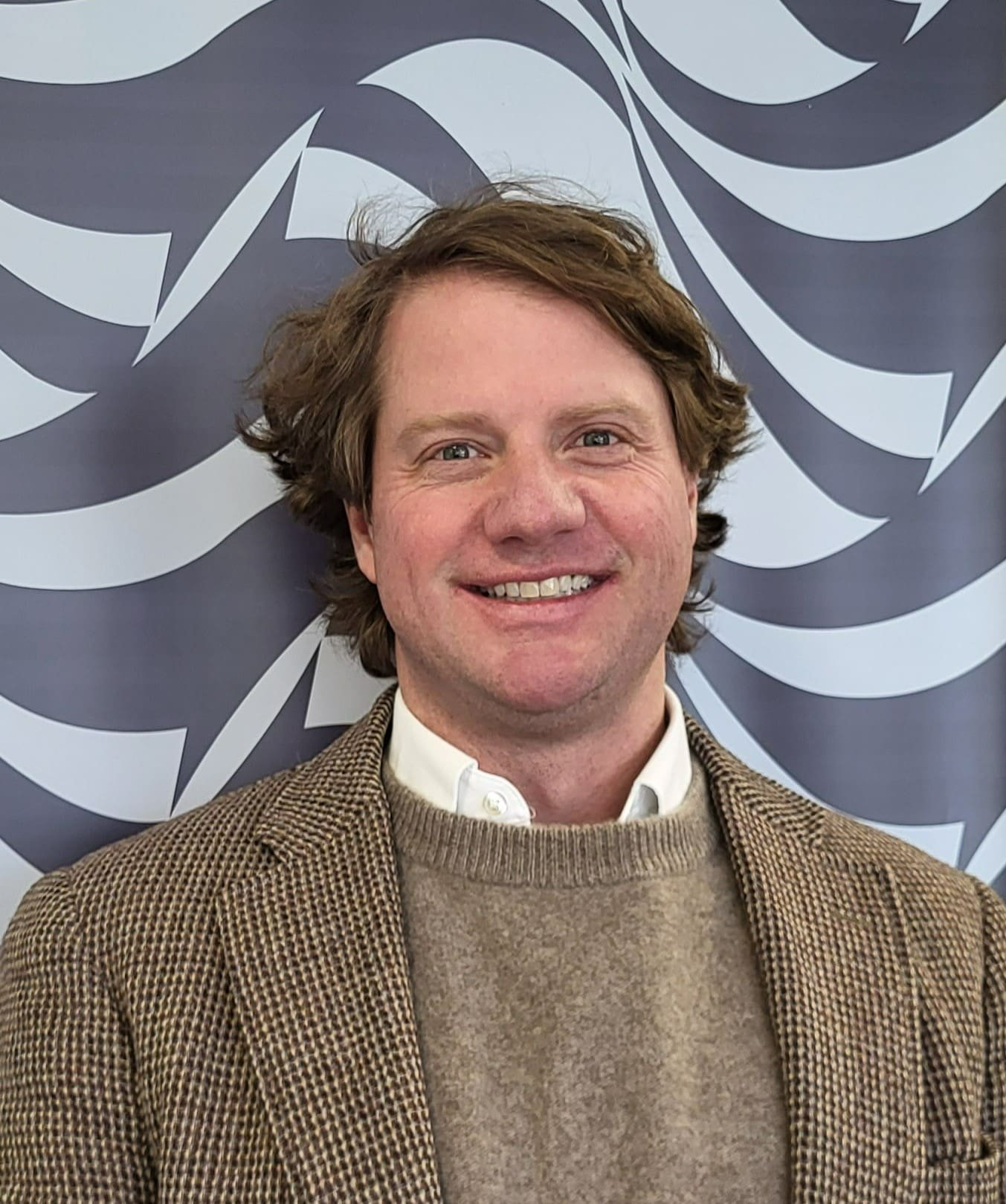 A smiling man with brown hair wearing a brown and beige houndstooth blazer, a white shirt, and a beige sweater, standing in front of a blue and white patterned backdrop.