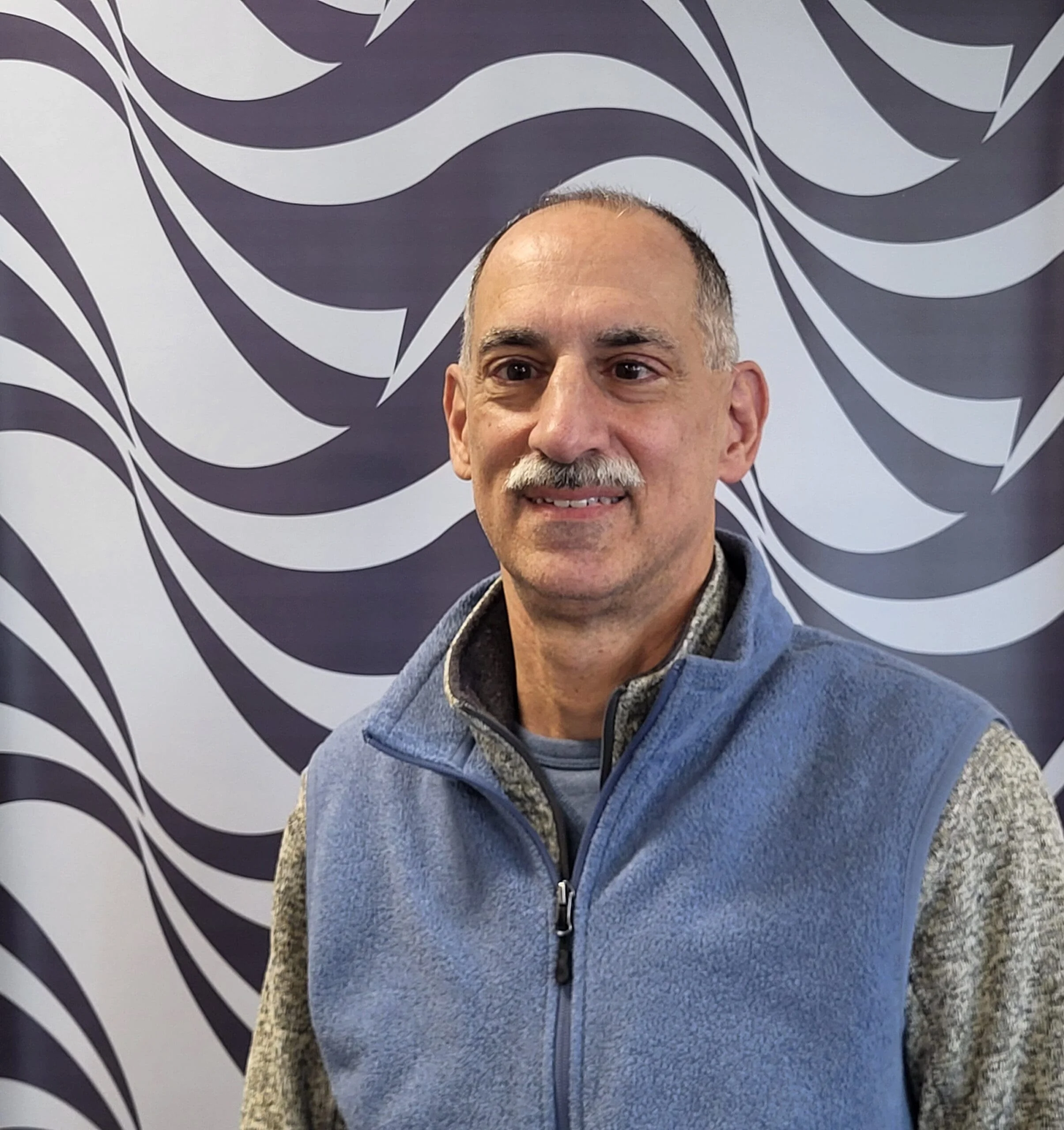 A middle-aged man with a mustache, wearing a blue zip-up vest over a gray and black patterned sweater, standing in front of a black and white wave-like patterned background.