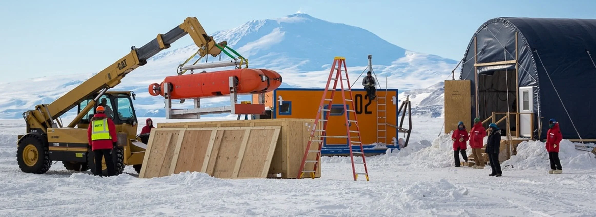 Research team setting up equipment on snow-covered landscape, with mountains in the background, using a crane to lift an orange cylinder; some workers are standing, others are on a ladder near a dark tent.