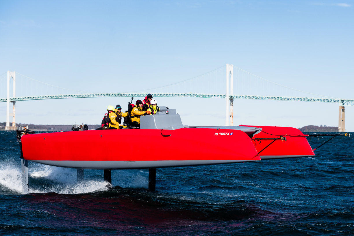 A red and gray seaplane flying over water with a bridge in the background, carrying several people dressed in yellow and black jackets and helmets.
