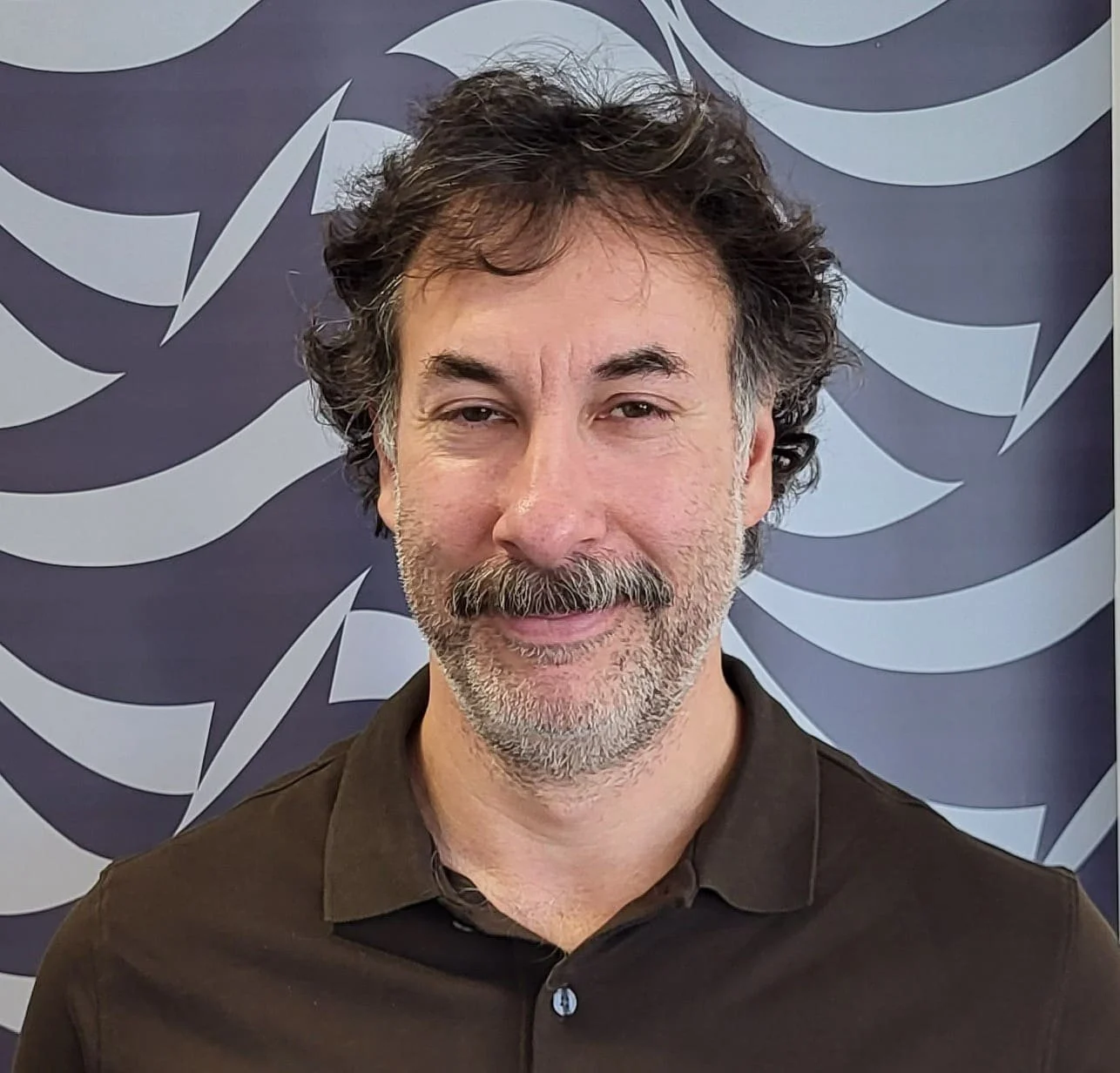 A middle-aged man with bushy dark hair and a beard, wearing a dark button-up shirt, standing in front of a dark background with abstract light-gray and dark-gray curved shapes.