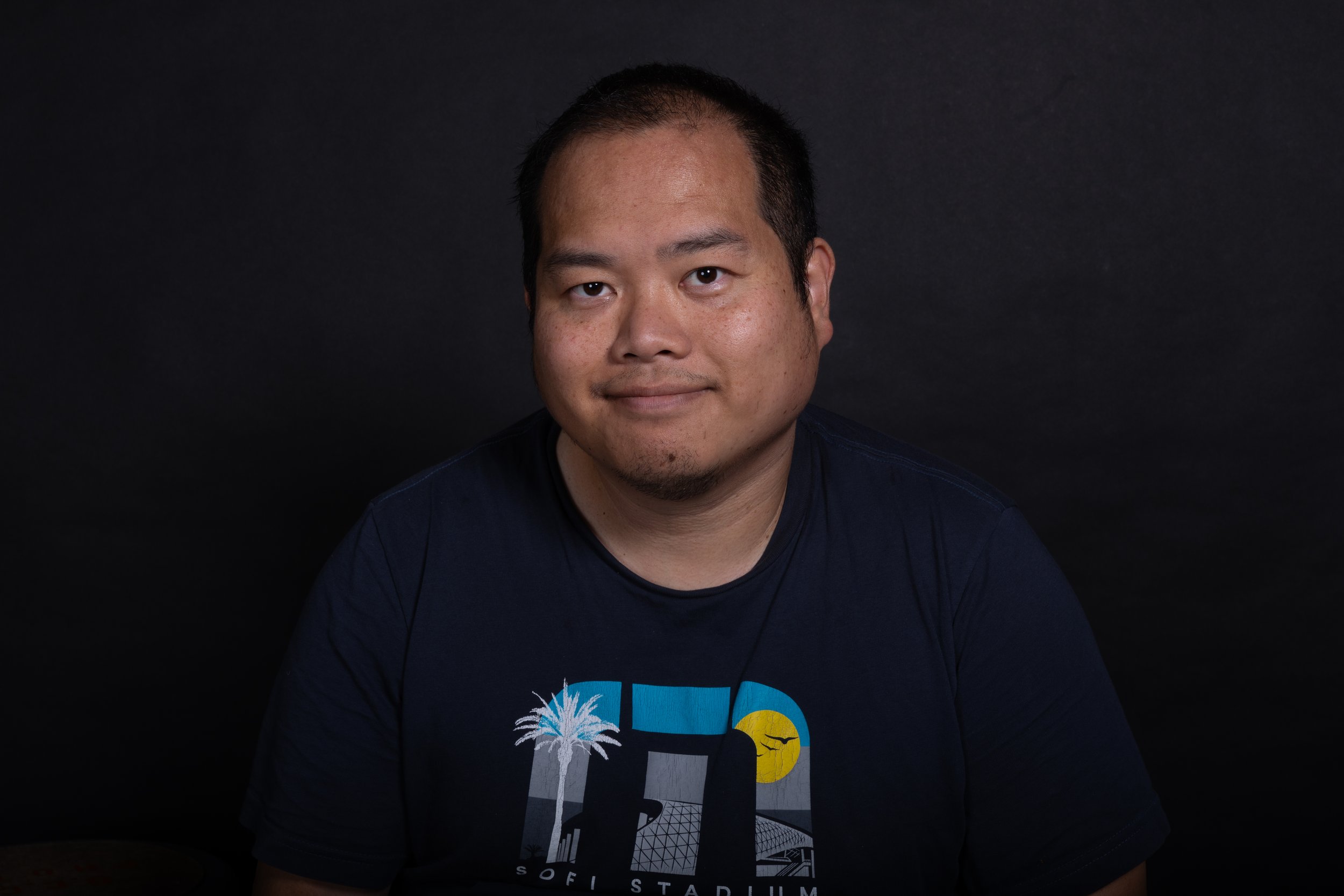 A man with short dark hair and a slight beard smiling in front of a black background. He is wearing a dark T-shirt with a colorful graphic design featuring palm trees, a sun, and geometric patterns.