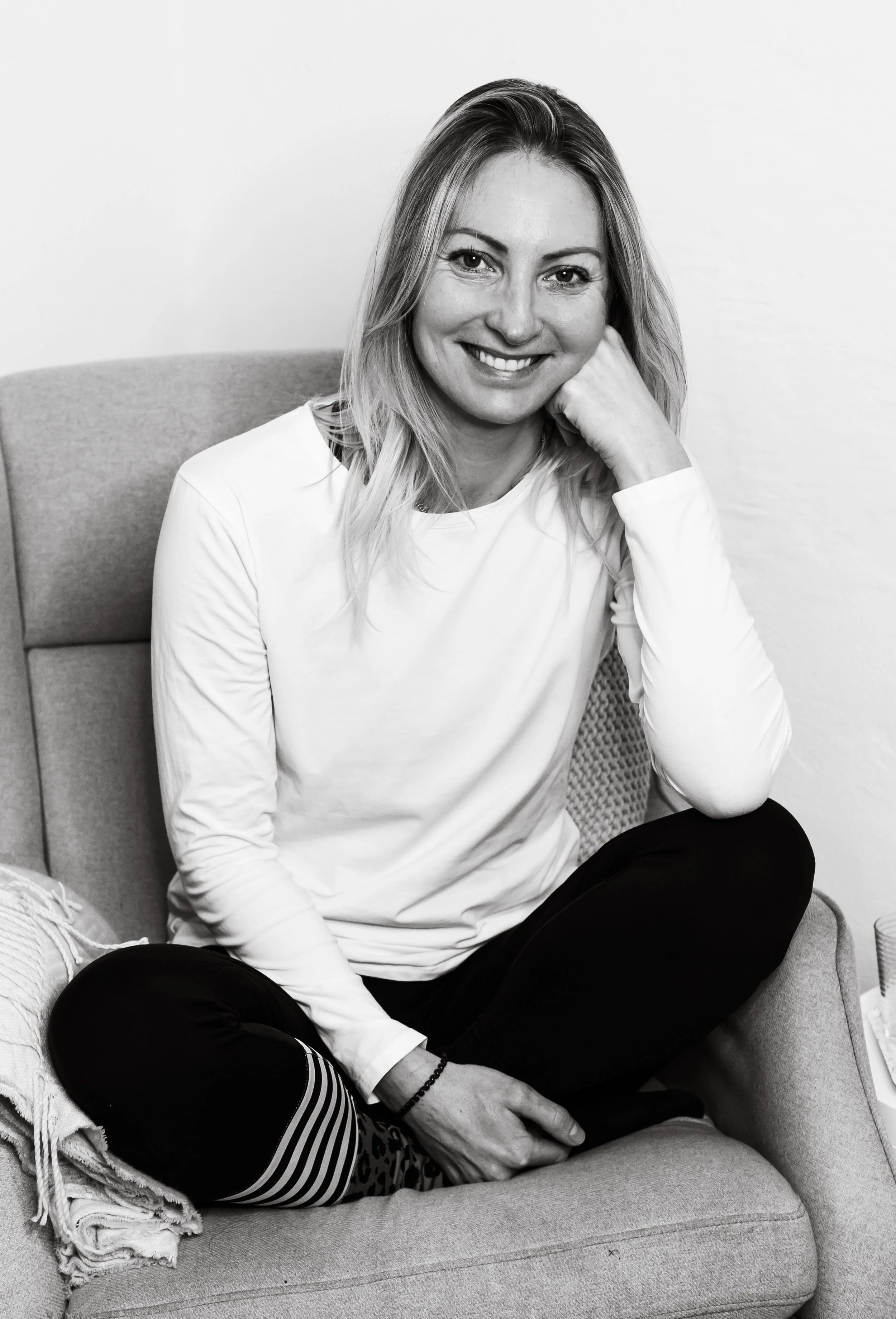 Schwarz-Weiß-Foto einer lächelnden Frau, die bequem auf einem Sofa sitzt, mit Hand am Kopf, in einem weißen Longsleeve. Sie trägt schwarze Hosen und blickt direkt in die Kamera.