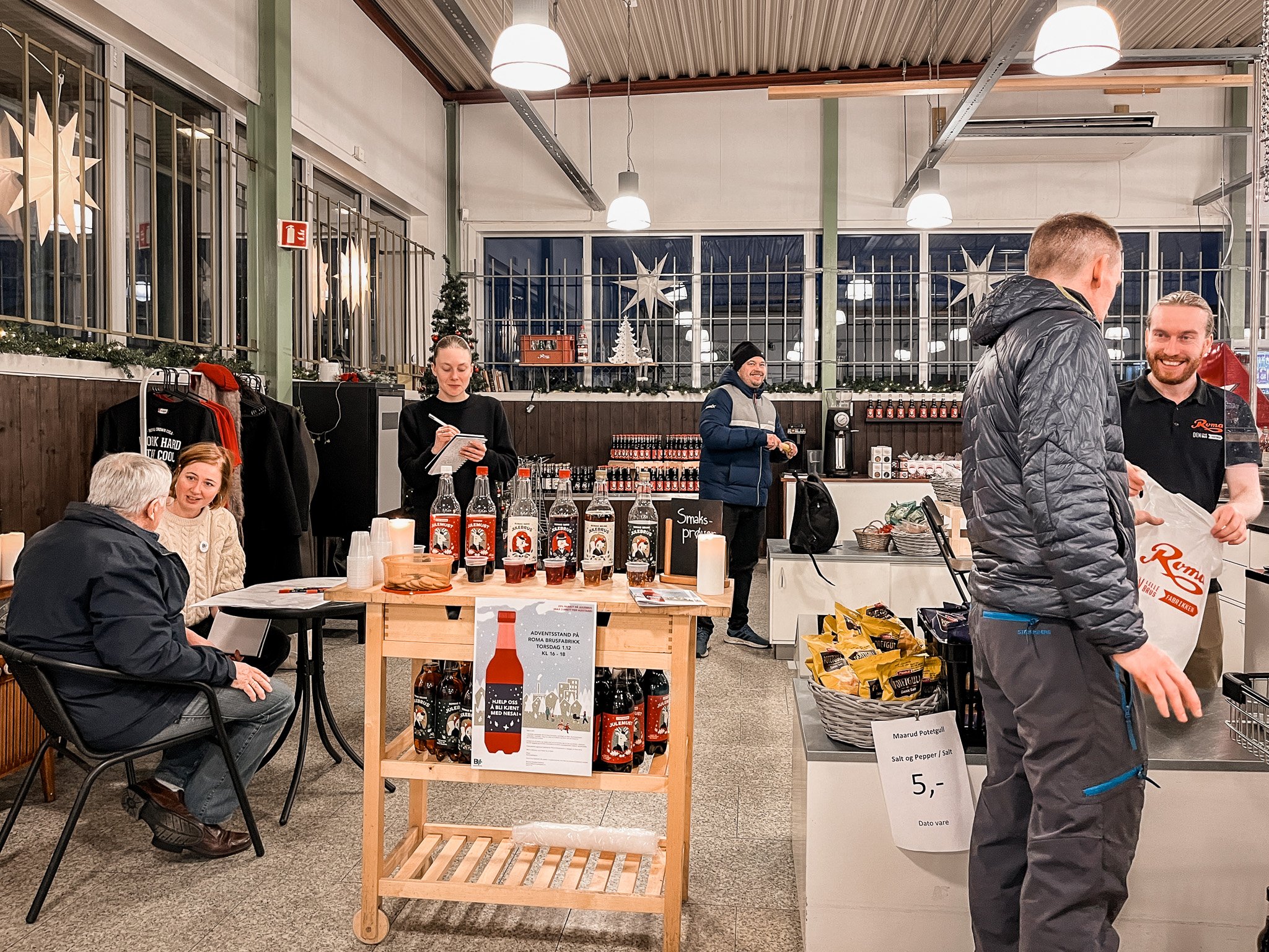 People inside a store decorated for Christmas, with Christmas lights and stars in the window. Two men at the counter exchanging items, one smiling, and a woman taking notes in the background. A table with bottles and a sign for sale, with customers seated nearby.