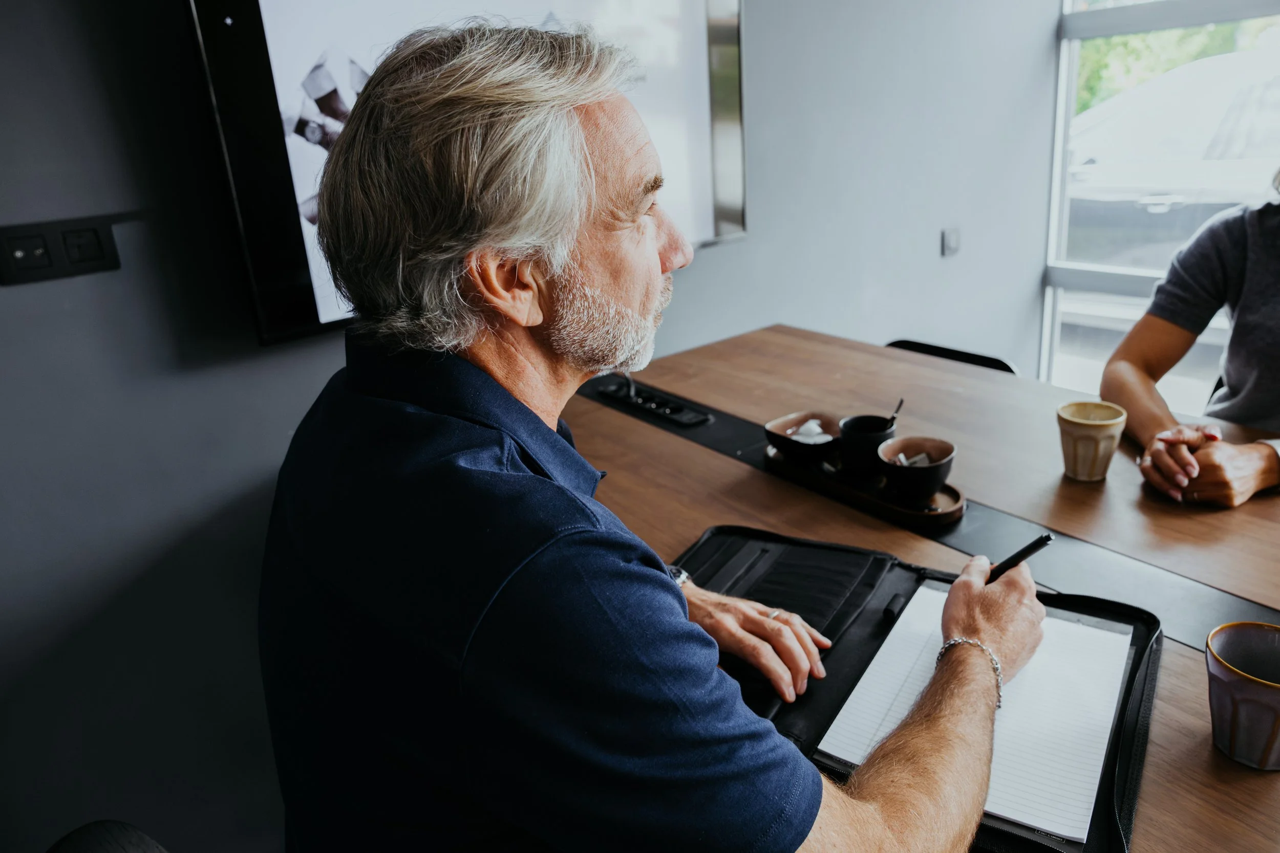 Een oudere man met grijs haar en een korte baard schrijft op een notitieblok tijdens een vergadering in een moderne kantoorruimte.