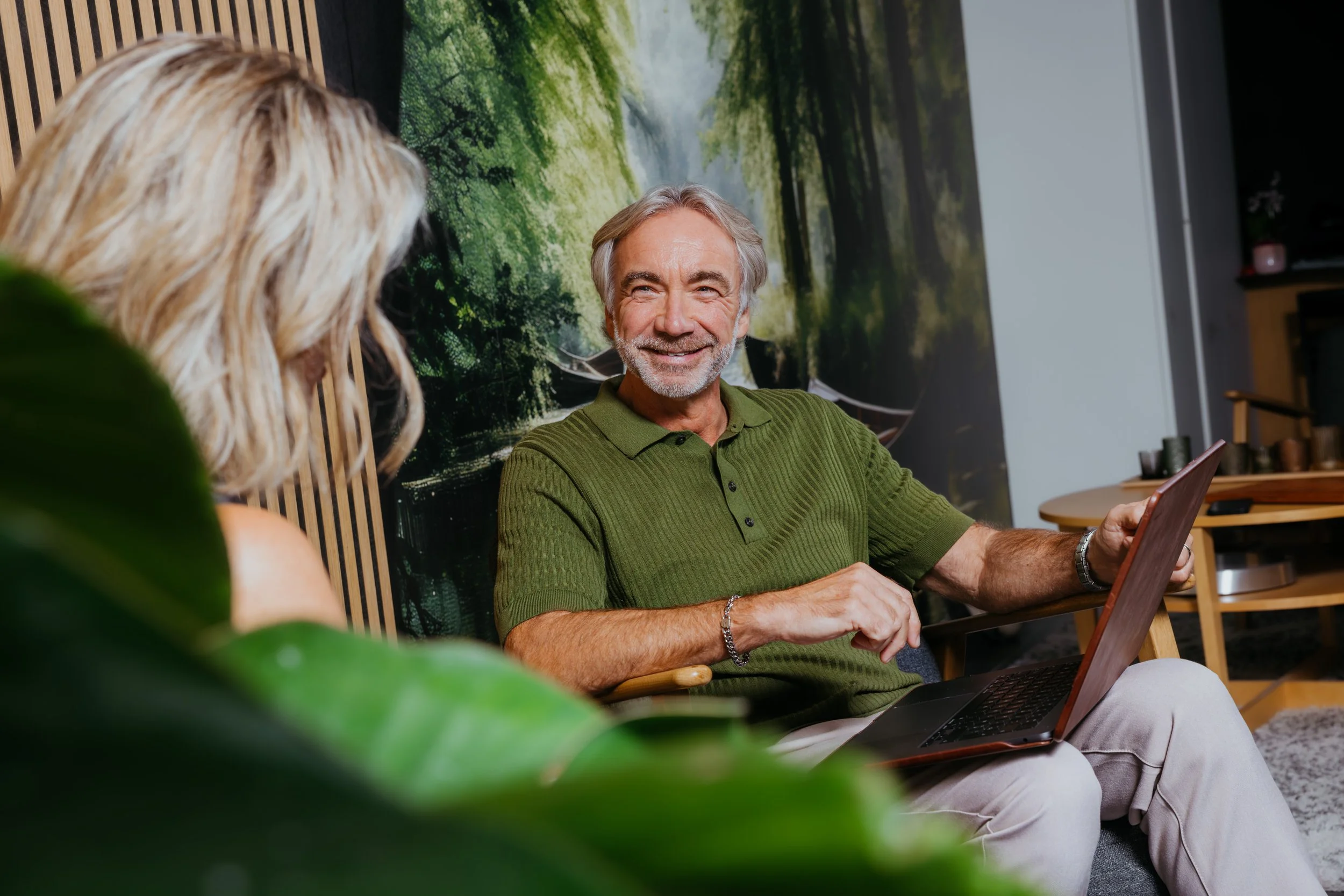Een oudere man met grijs haar en een baard, glimlachend, zit in een comfortabele stoel met een laptop op zijn schoot. Hij draagt een groene polo en een armband. Een vrouw met lang blond haar, van achteren zichtbaar, zit tegenover hem. Ze bevinden zich in een gezellige ruimte met een grote muurposter van een boslandschap op de achtergrond.