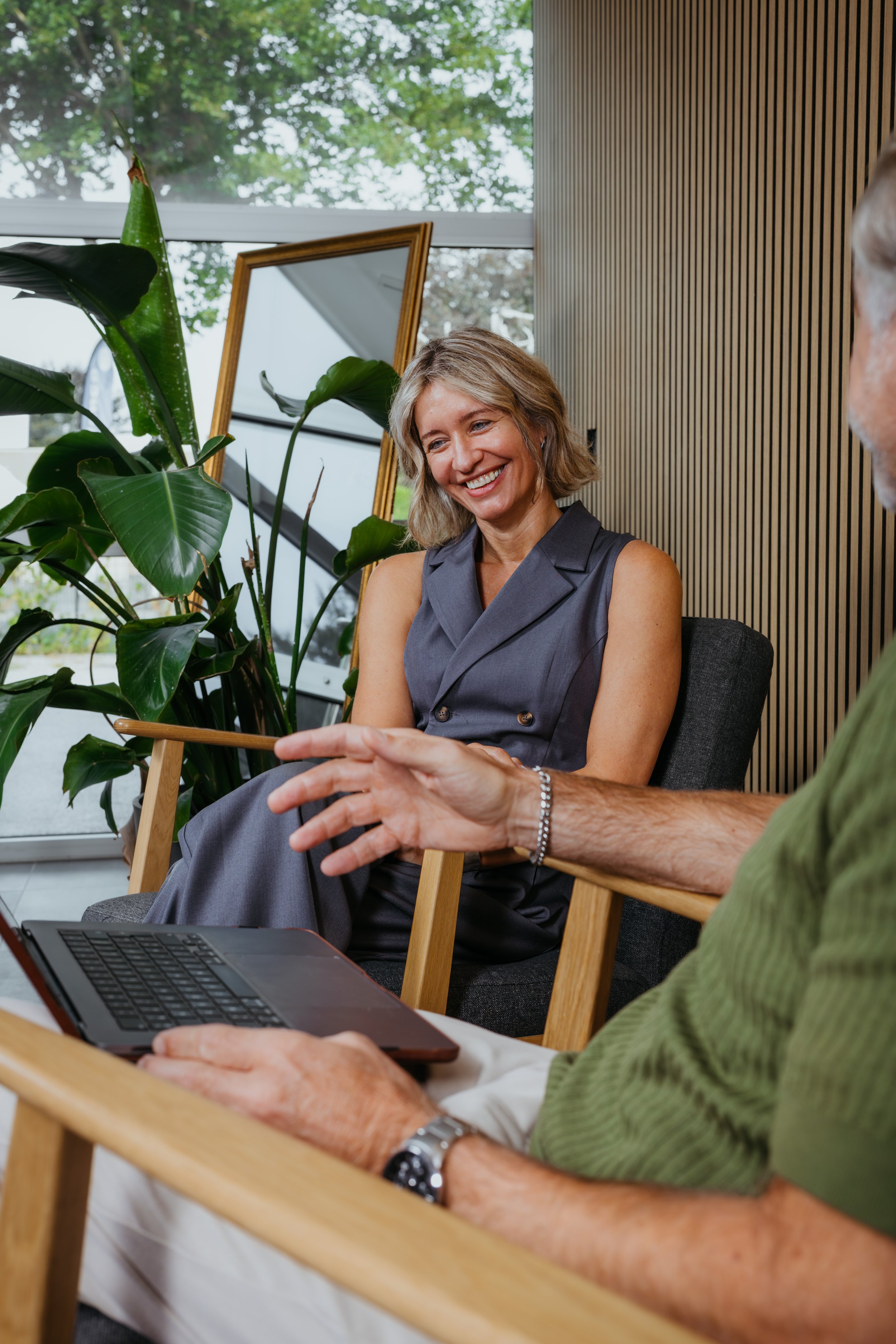 Twee mensen in gesprek in een moderne kamer met grote raampartij en groene planten, vrouw glimlacht terwijl ze mensen vertelt.