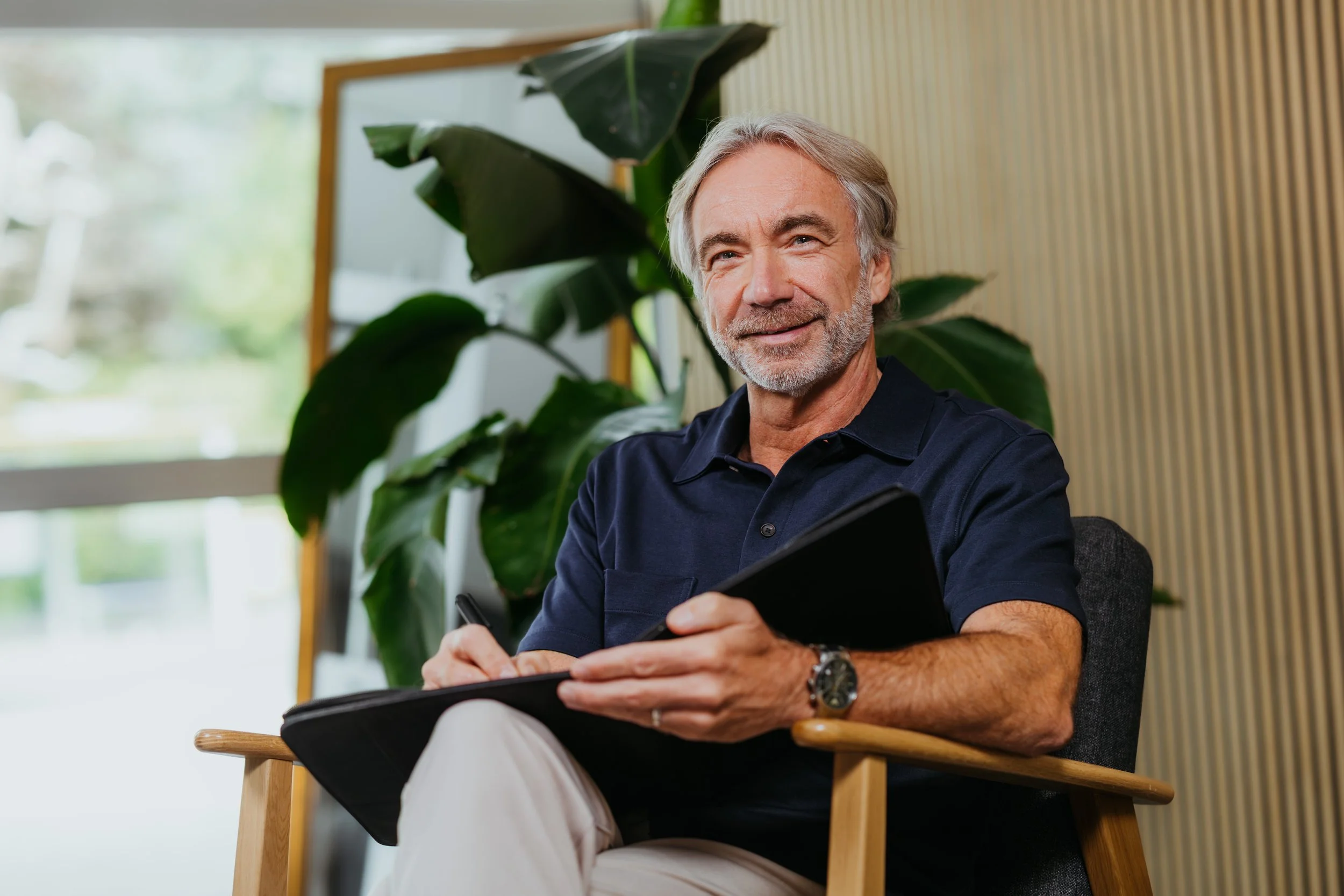 Een oudere man met grijs haar en baard zit op een stoel, houdt een tablet en een pen vast, in een kantoor of woonkamer met grote plant en raam.
